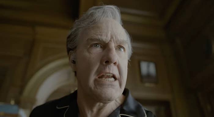 Man with intense expression, wearing a dark collared shirt with light piping, inside a wood-paneled room