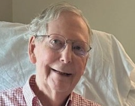 An elderly man with glasses smiles while seated. He is wearing a checkered shirt and has light hair
