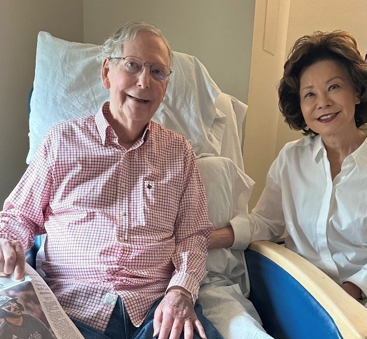 Two people seated, one in a checkered shirt holding a newspaper, the other in a white blouse. They appear relaxed and are smiling