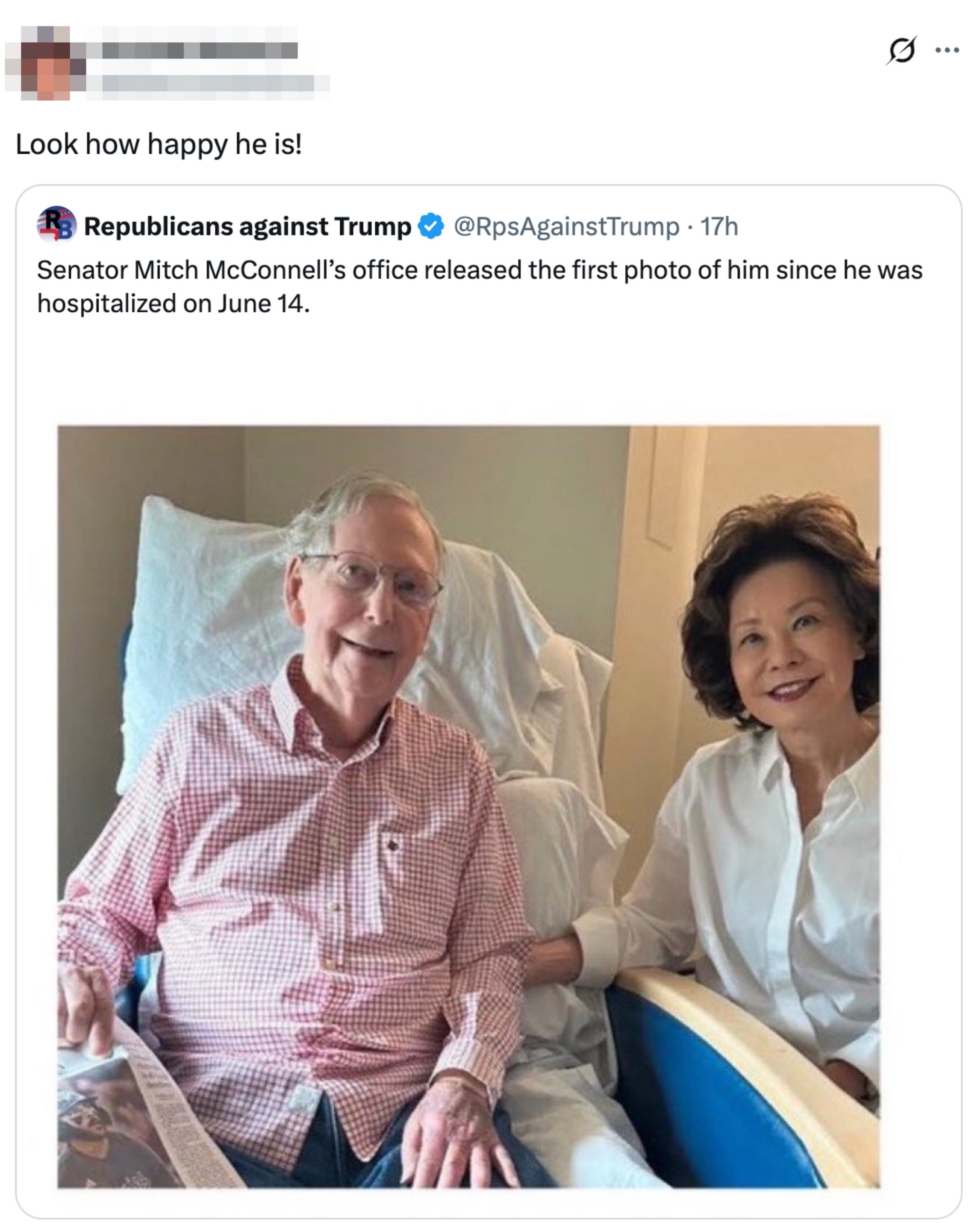 A man in a hospital setting sits in a chair while a woman in a blouse sits beside him, holding his arm. Both are smiling at the camera