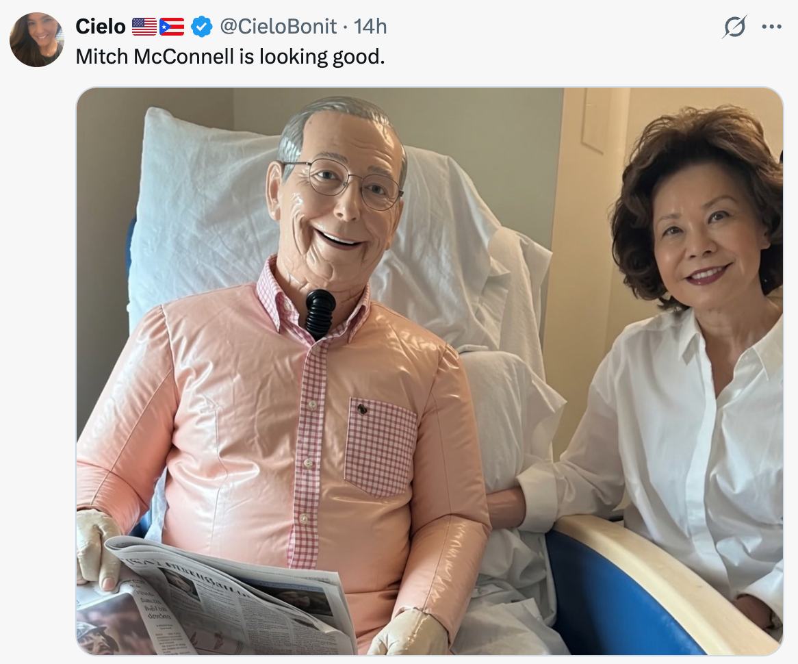 A person smiles and holds hands with a figure resembling a ventriloquist dummy dressed in a casual shirt, sitting on a chair with a newspaper