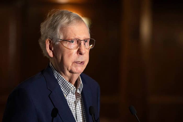 An older man in glasses and a checkered shirt speaks at a podium, captured in a professional setting