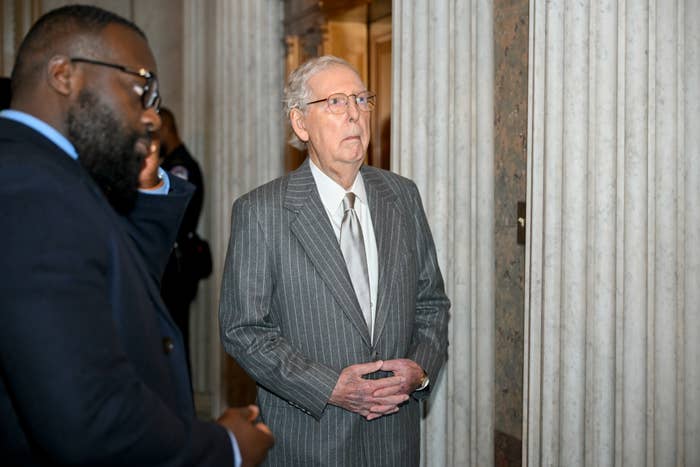An elderly man in a pinstripe suit stands indoors, surrounded by others