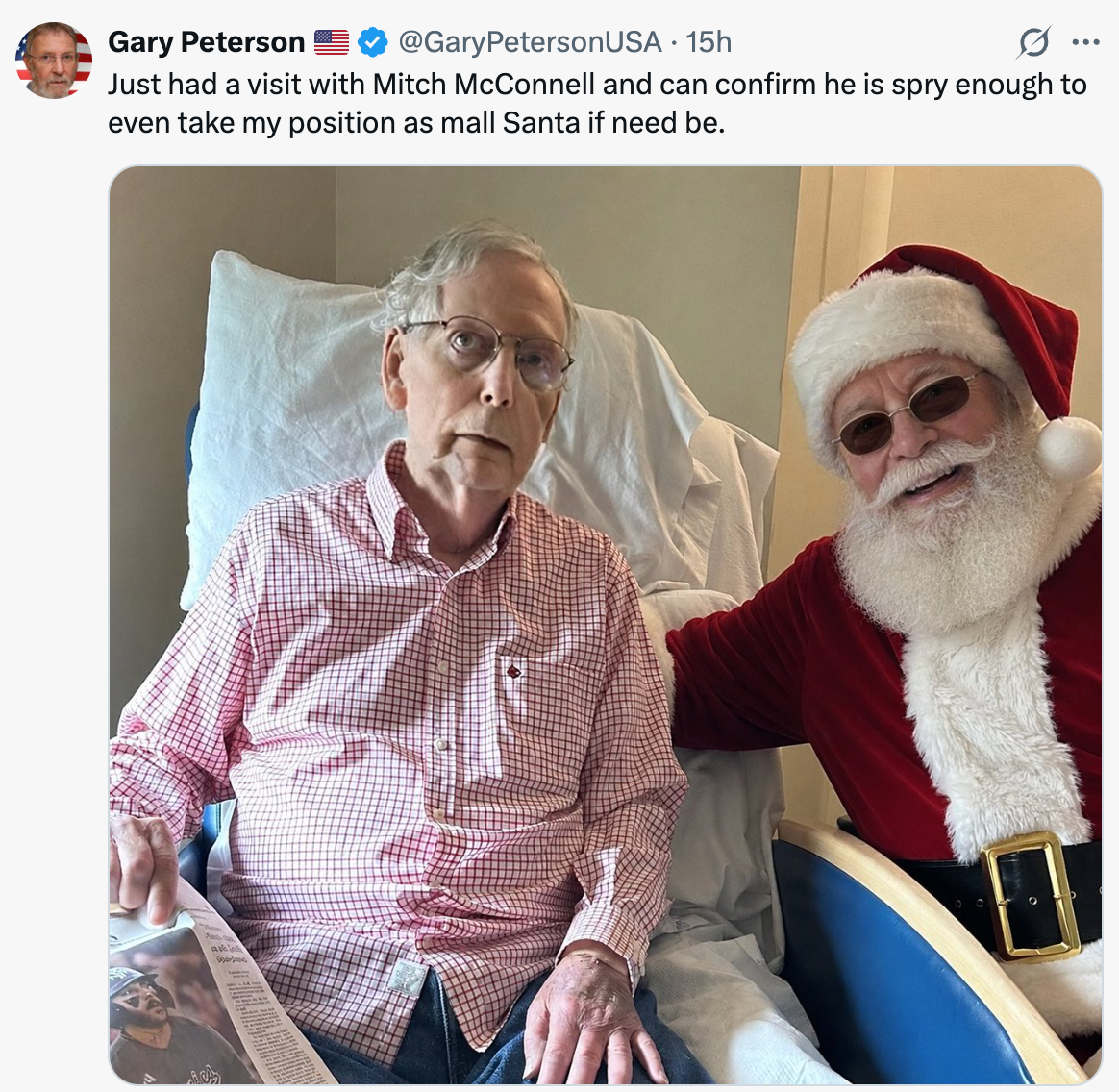 An elderly man in a casual shirt sits in a chair, while a person dressed as Santa Claus sits beside him, smiling and resting a hand on his shoulder