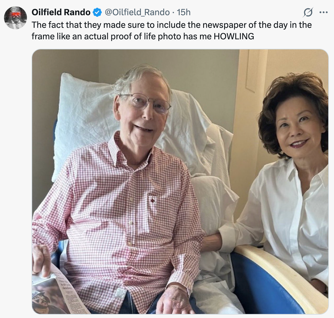 Two people smile while seated; one holds a newspaper. The tweet above humorously comments on the inclusion of the newspaper in the photo