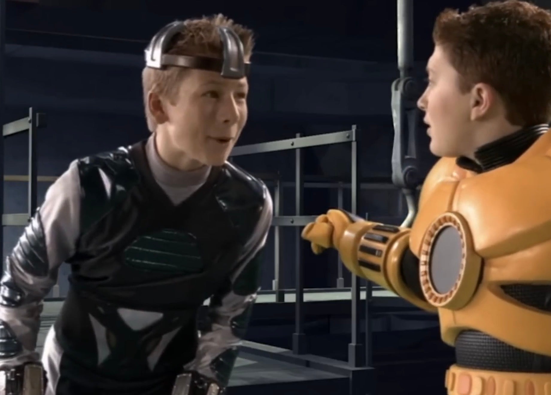 Young Glen Powell and Daryl Sabara in futuristic costumes having a lively conversation in a sci-fi setting