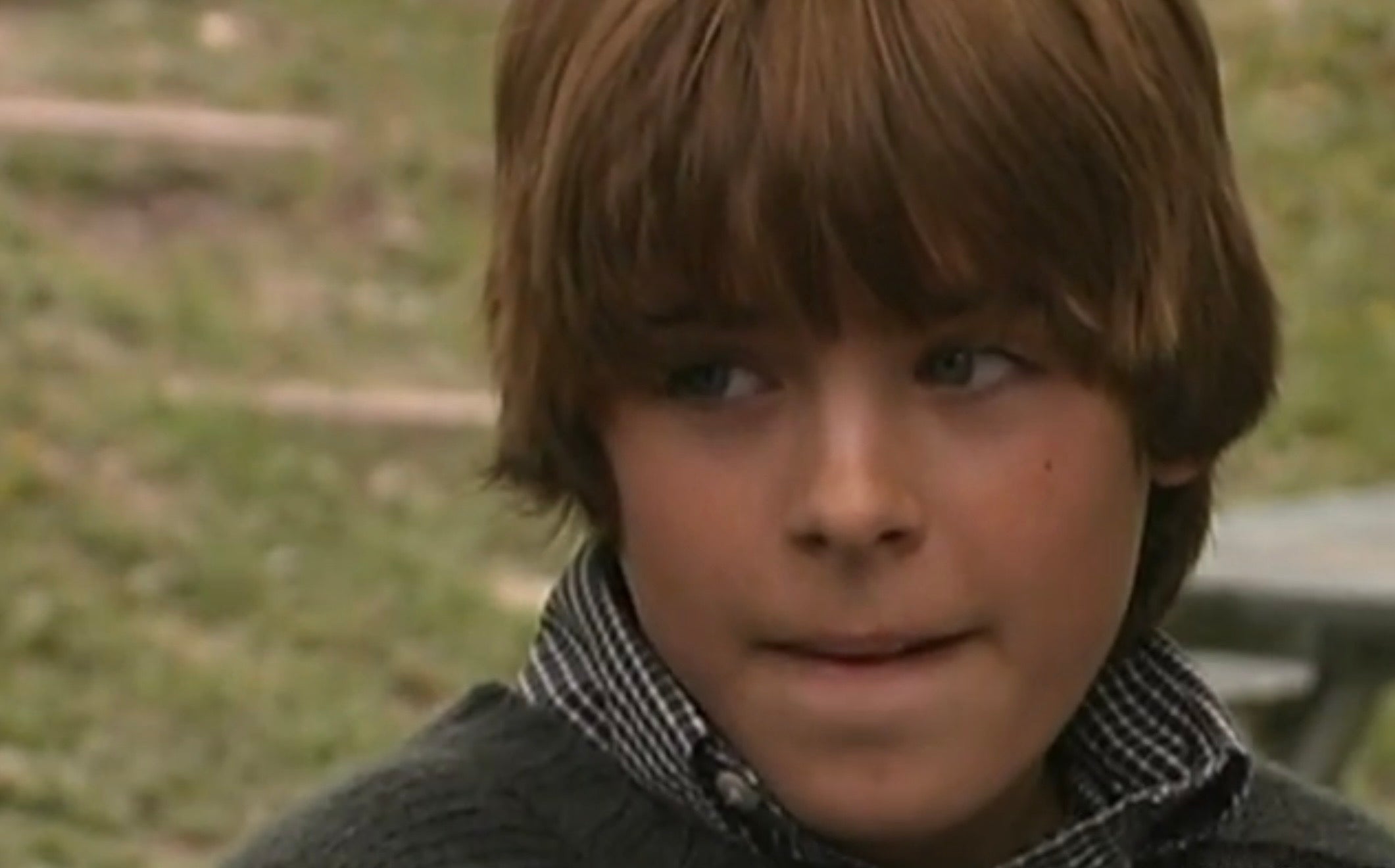 Young Zac Efron outdoors, looking to the side with a thoughtful expression, wearing a checked shirt under a sweater
