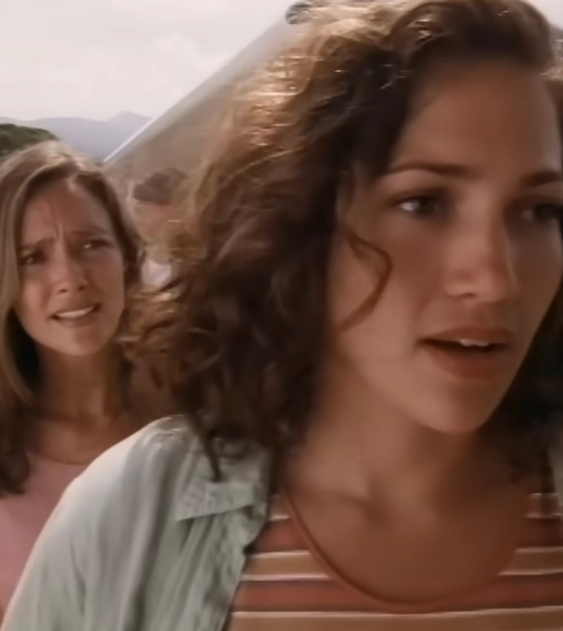 Jennifer Lopez with curly hair stands outdoors near an aircraft, wearing a striped shirt and open jacket, appearing thoughtful