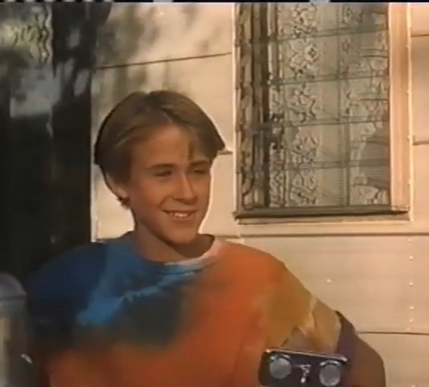 Young Ryan Gosling smiling, wearing a tie-dye t-shirt, standing outside near a window