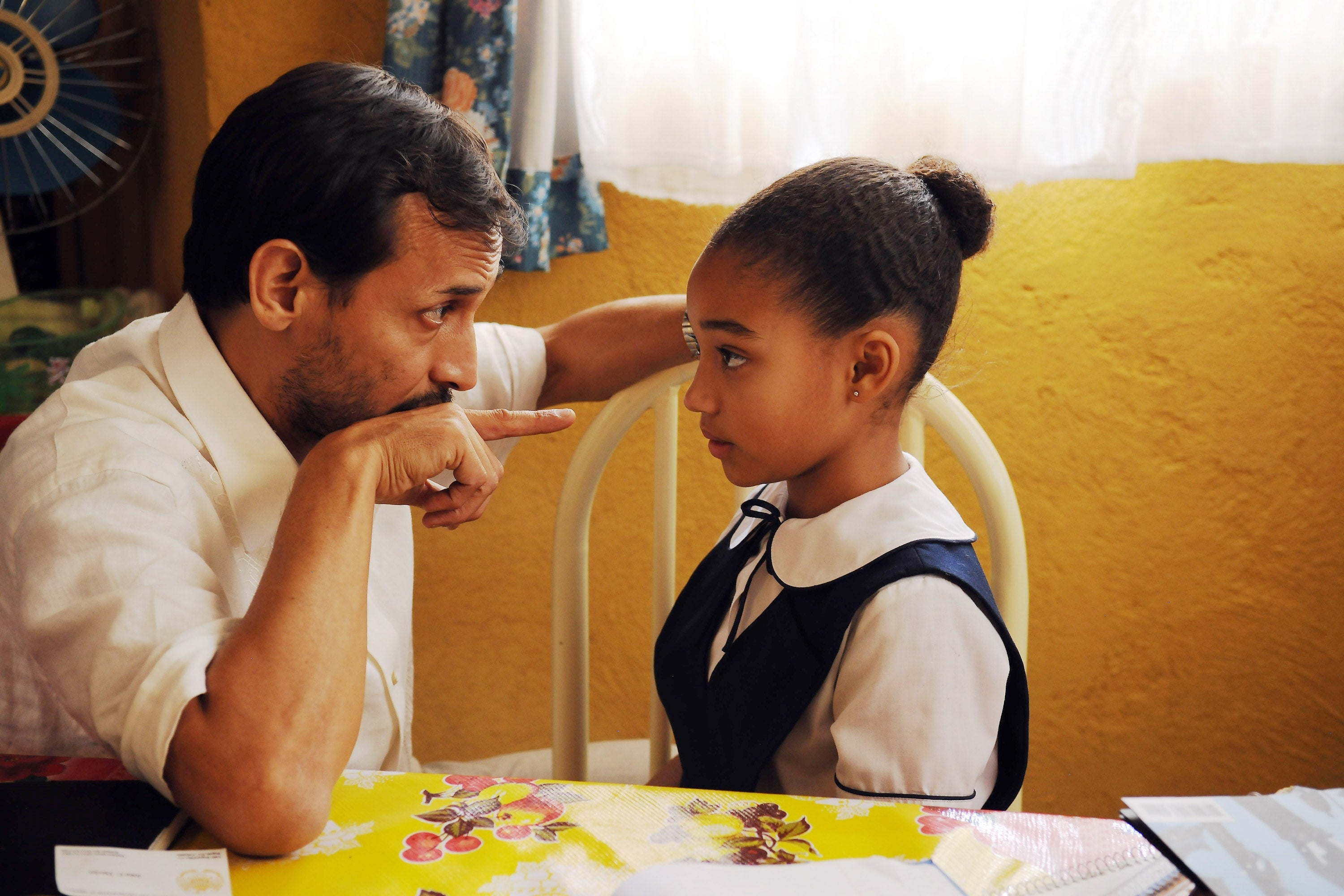 Jesse Borrego sitting at a table, intently talking to young Amandla Stenberg  in a school uniform, who is listening seriously