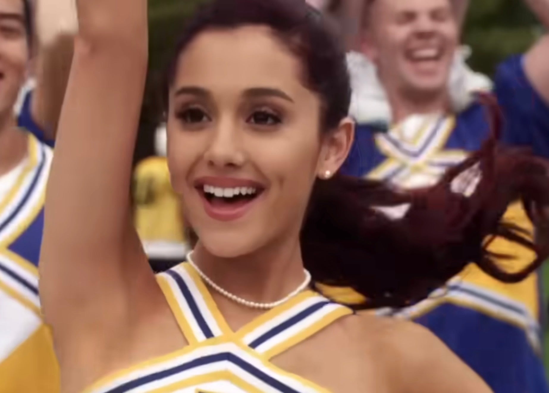 Ariana Grande dressed as a cheerleader with a crisscrossed neckline smiles energetically, surrounded by others in similar uniforms