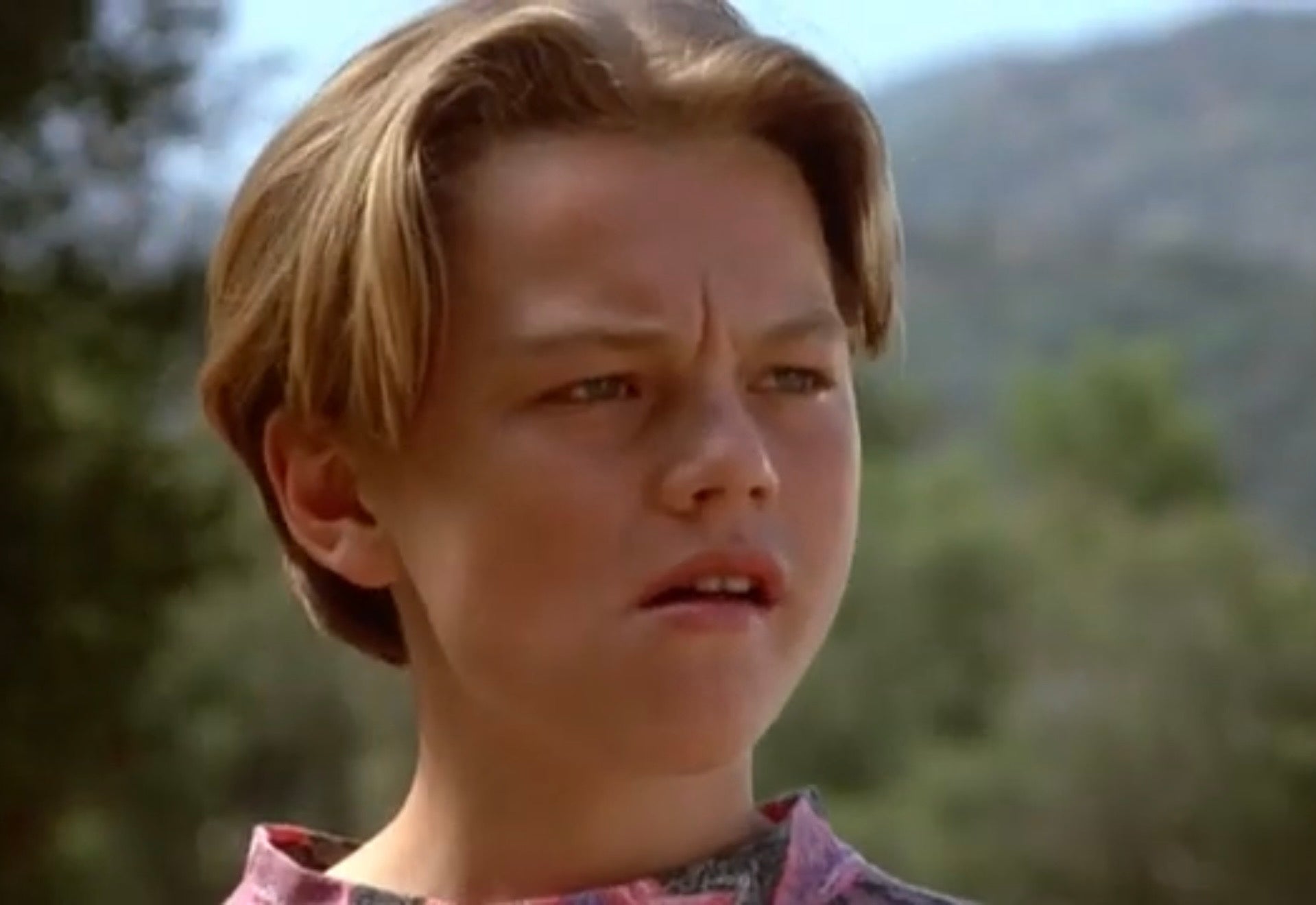 A young Leonardo DiCaprio with short hair appears thoughtful and slightly concerned, outdoors with trees in the background