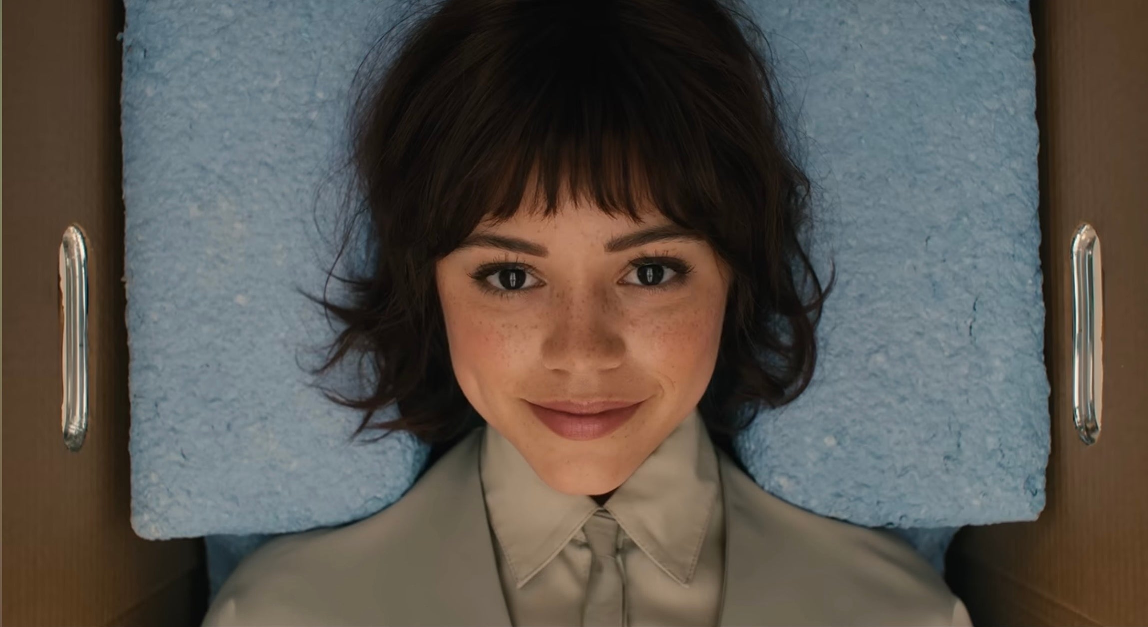 Jenna Ortega with short hair smiles while lying on a textured surface, wearing a collared shirt