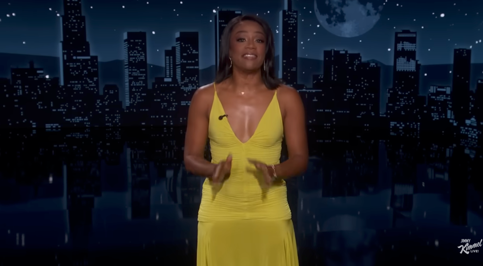 A pistillate   stands connected  a nighttime cityscape signifier    wearing an elegant, sleeveless formal  with a heavy  neckline, speaking to an audience