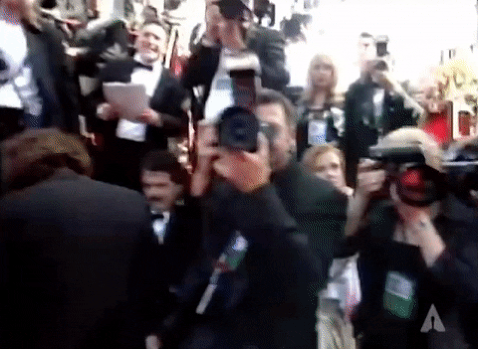 Photographers take pictures on a bustling red carpet at an event, with people in formal attire in the background