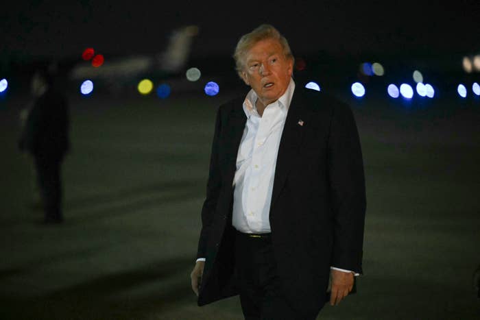 A person in formal attire stands on an airstrip at night, surrounded by small lights and a plane in the background