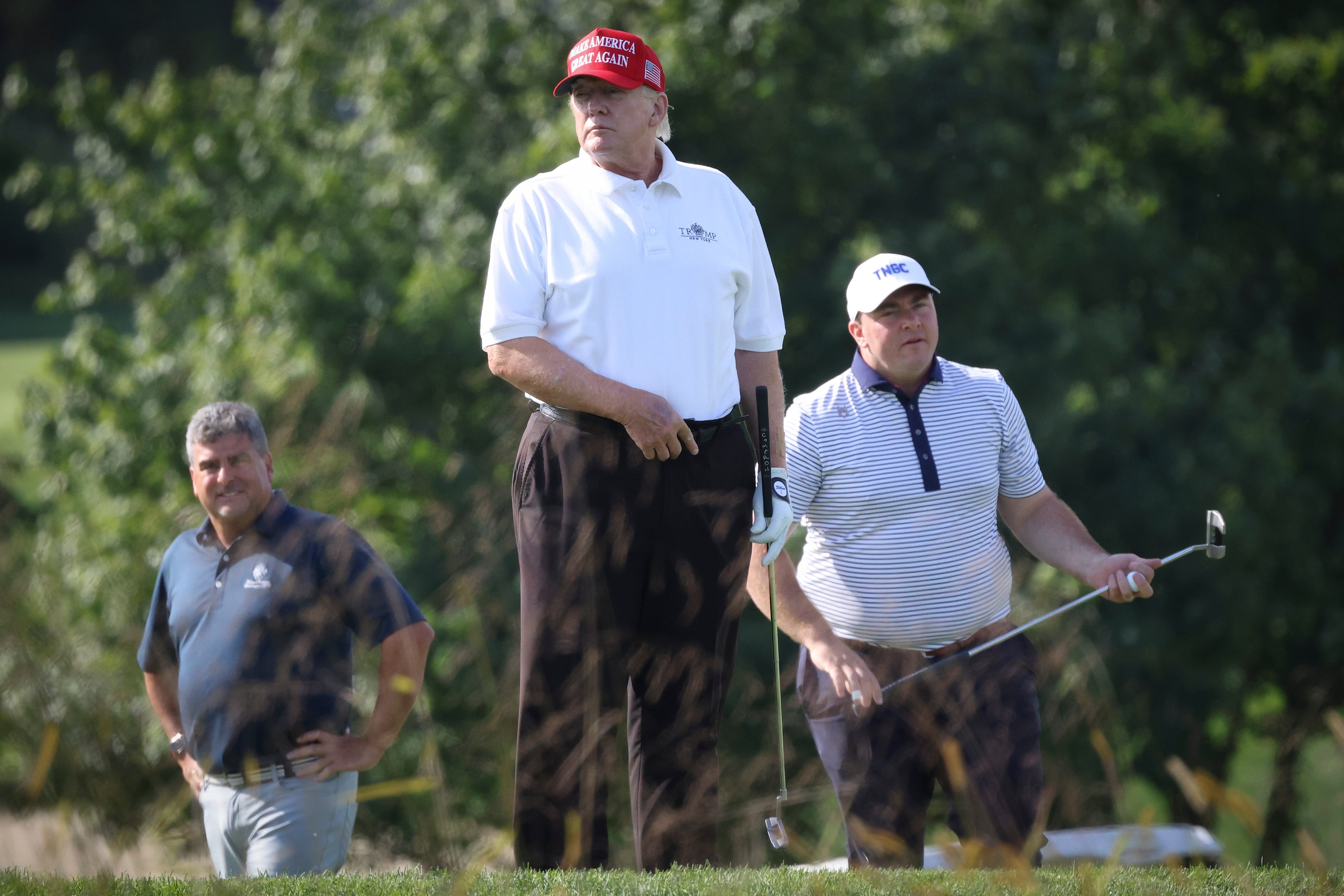 Tre personer er på en golfbane; en svinger en golfkølle, en har på seg en caps og en annen ser på. Trær er i bakgrunnen