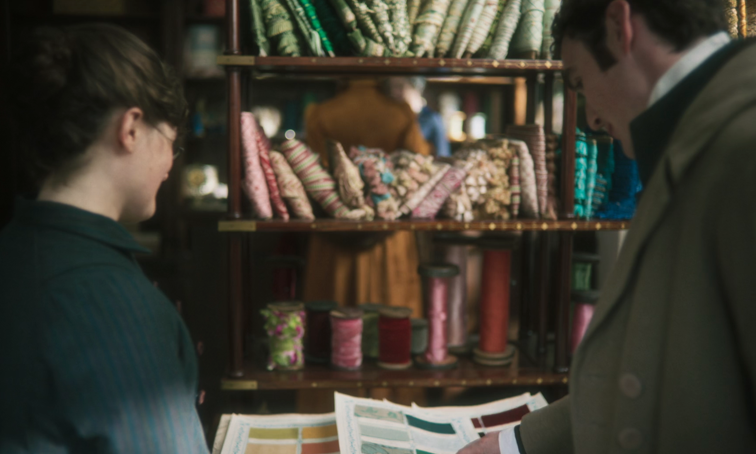 Two people in historical clothing examine fabric samples in a store with shelves filled with various patterned fabrics