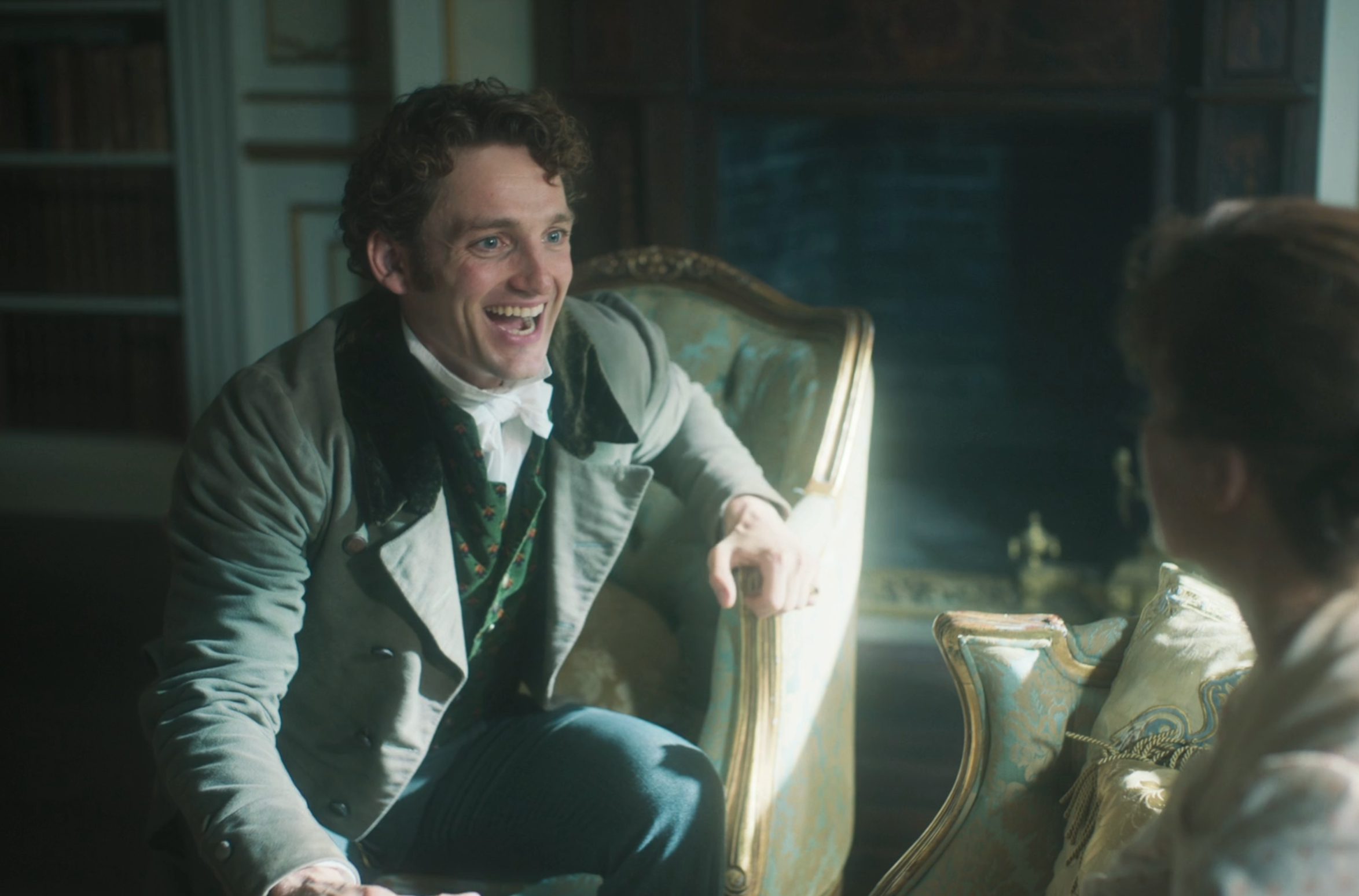 Two people in elegant period attire sit in a lavish room, talking and smiling