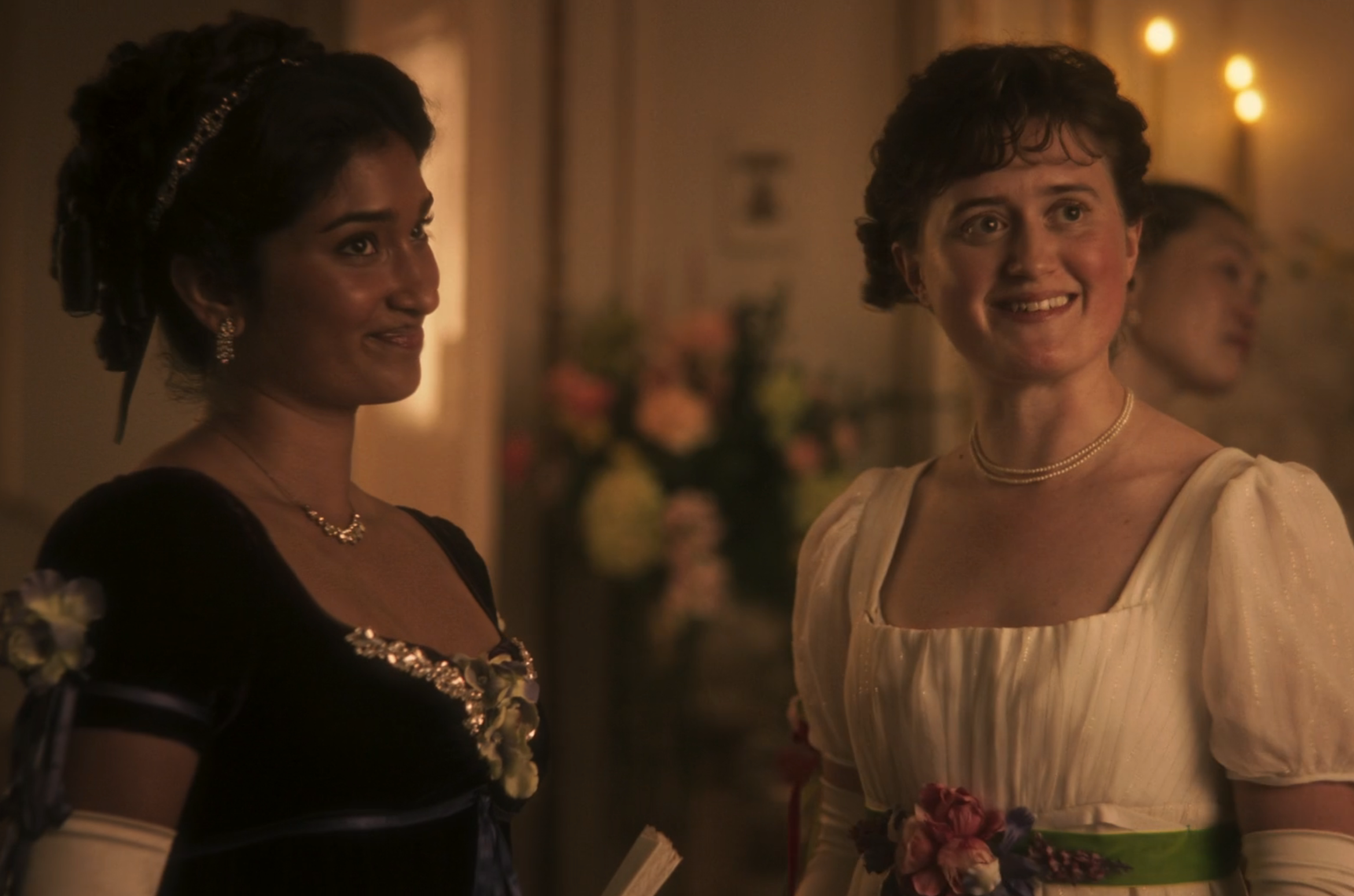 Two women in period-style gowns smile at an event. One wears a dark dress with ornate details, the other a light dress with floral accents