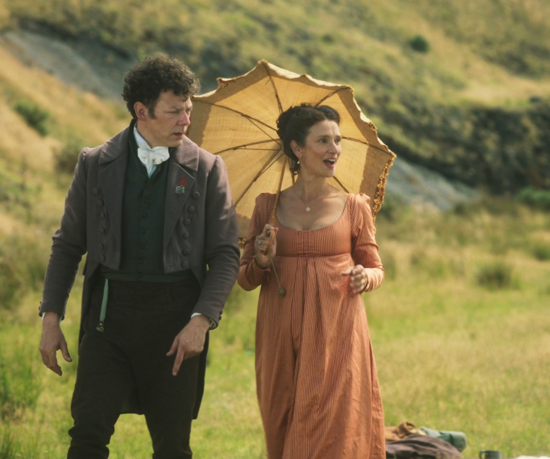 Two people in historical attire, one holding a parasol, stand in a grassy landscape. The style suggests a period setting, possibly a film or series scene