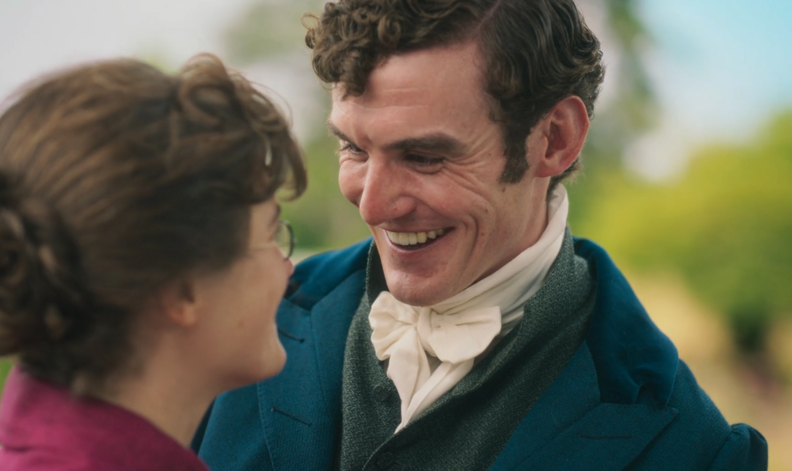 A man and a woman in period attire share a joyful moment outdoors, smiling at each other warmly
