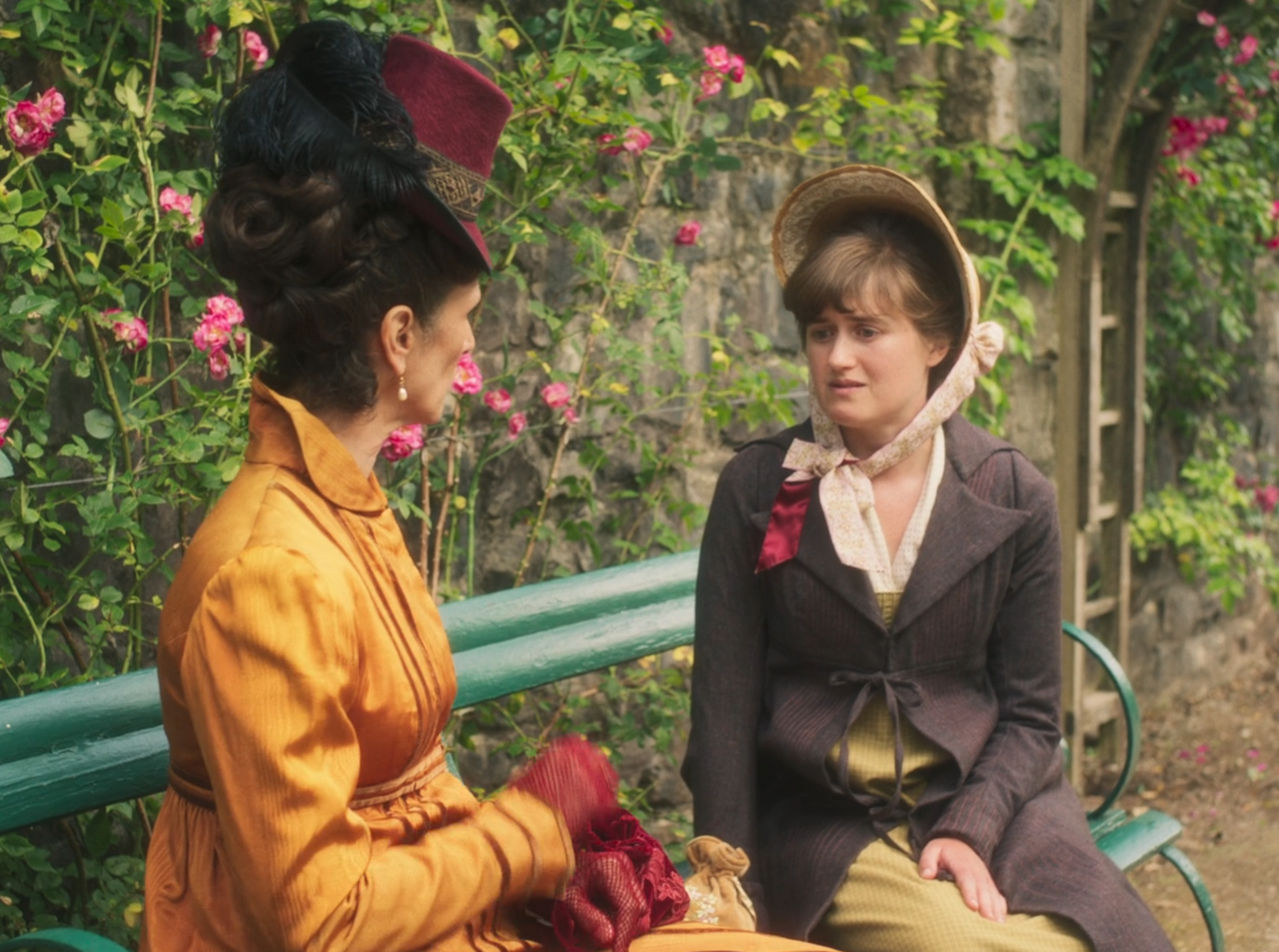 Two women in Victorian-era attire sit on a bench in a garden. One wears a top hat and the other a bonnet