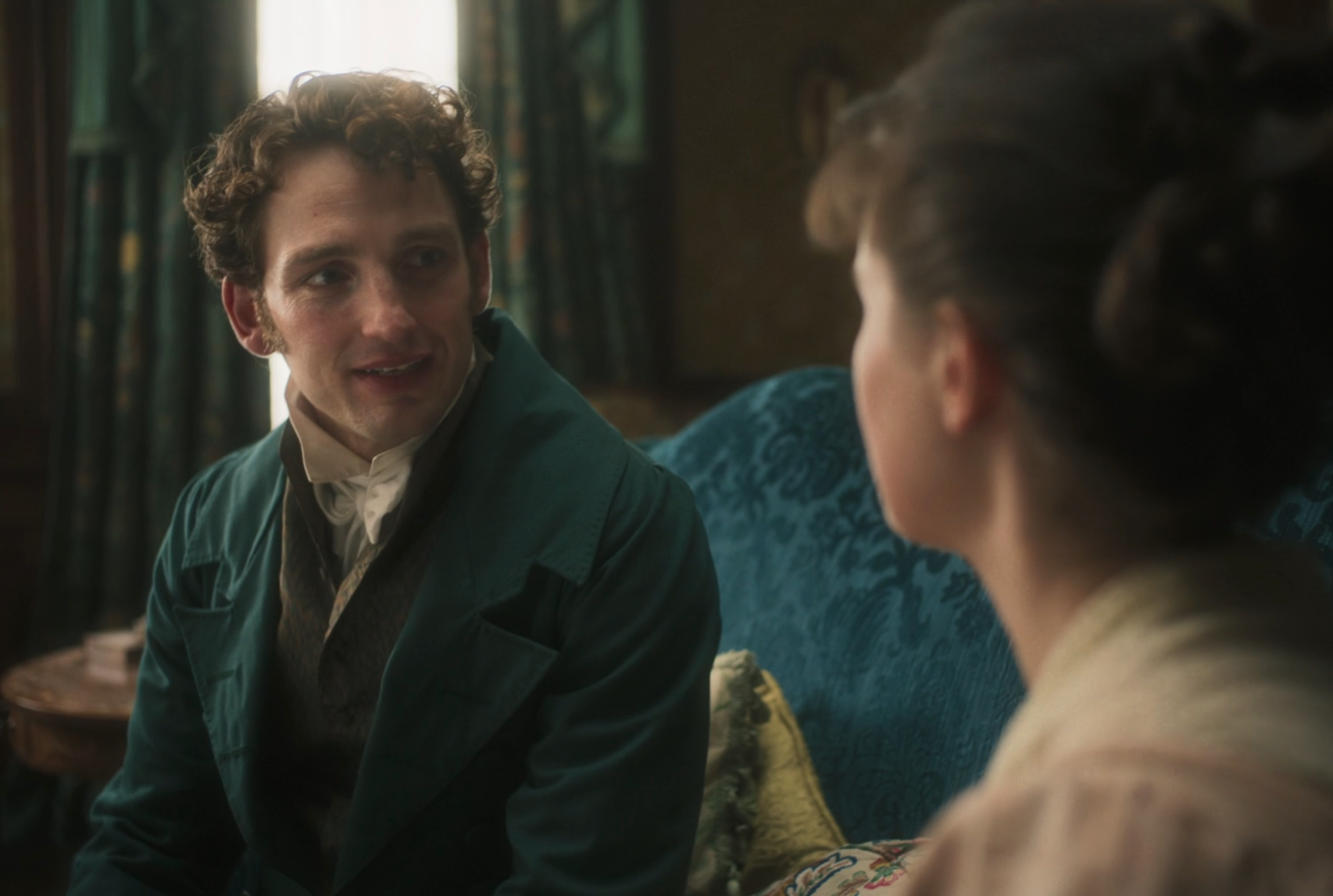 A man and woman in period clothing sit on a vintage couch in a historic room, engaged in conversation