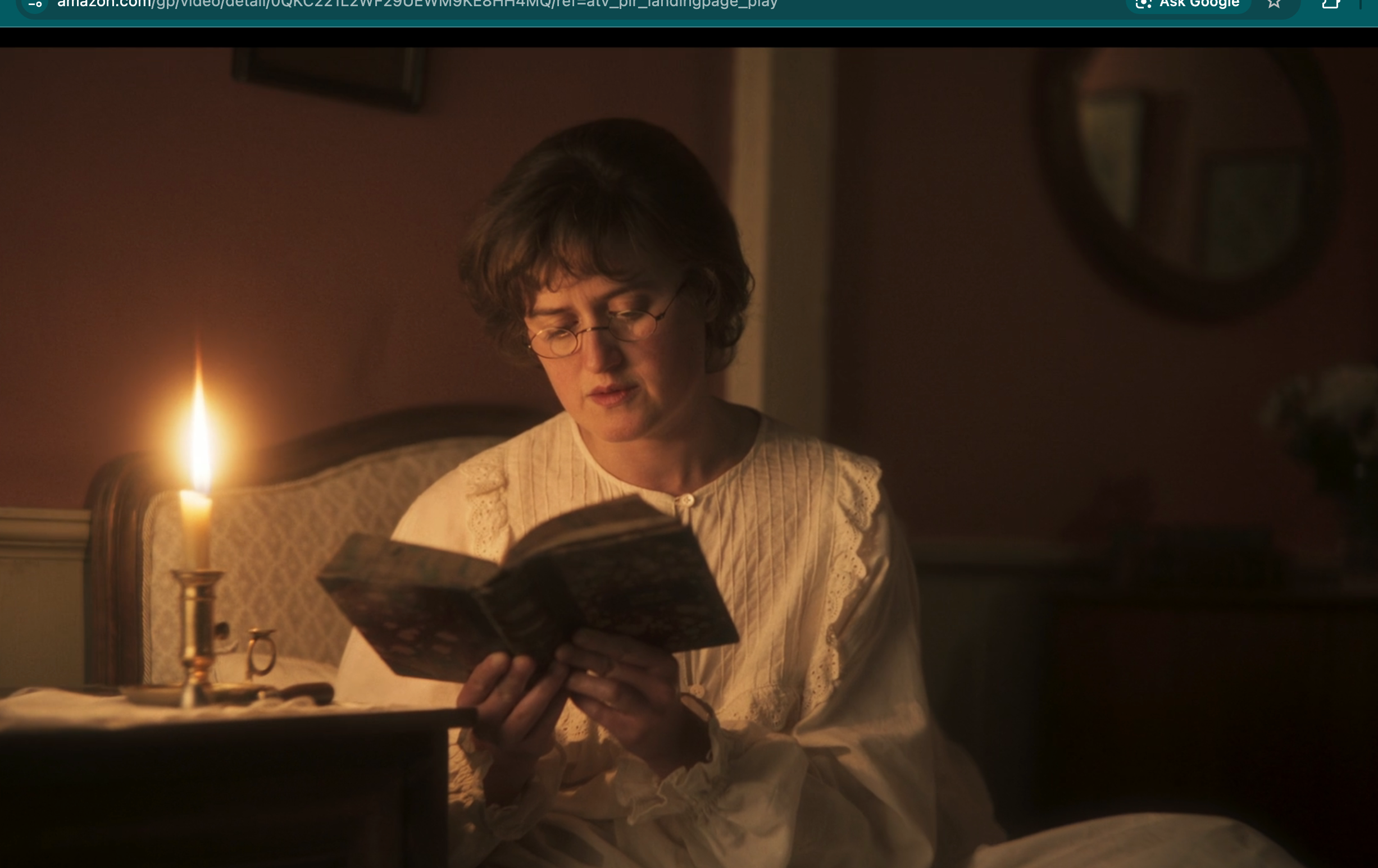 A person in period clothing reads a book by candlelight, seated in a dimly lit room