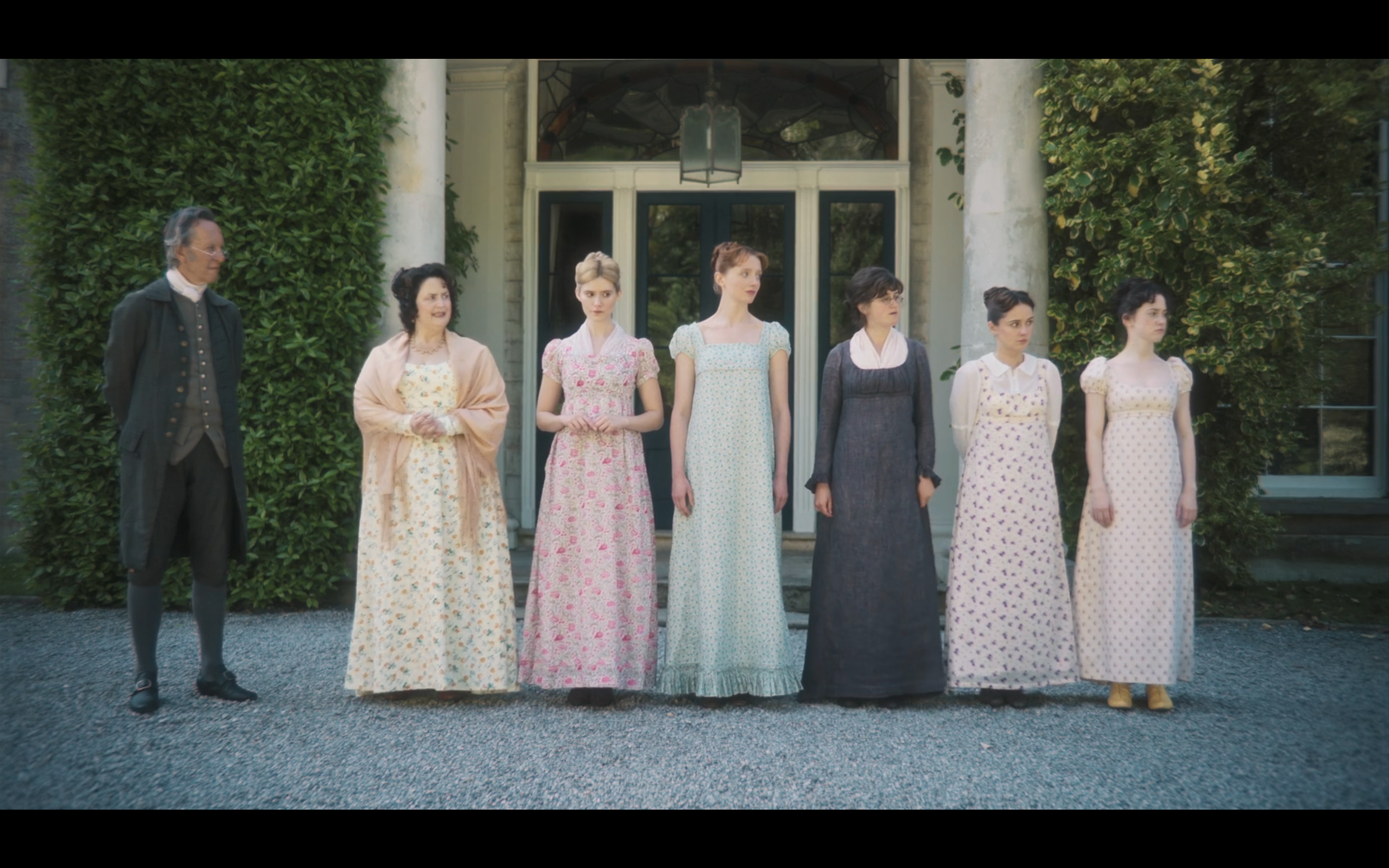 A group of six people stands outdoors in period costumes resembling Regency-era fashion, with women in long dresses and a man in a period suit