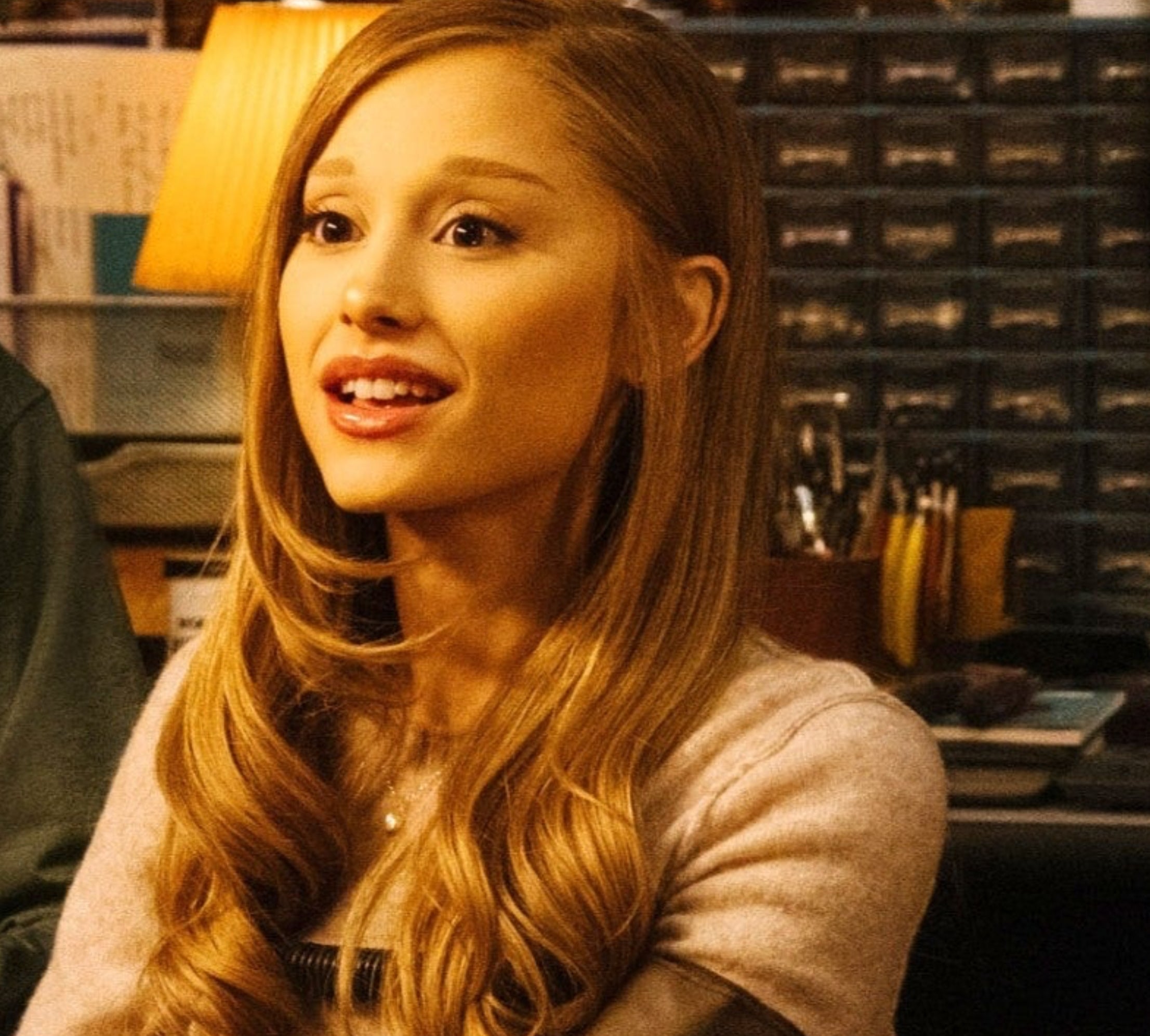 Ariana Grande with long hair smiles while sitting indoors, wearing a casual sweater. Background includes office supplies and a lit lamp