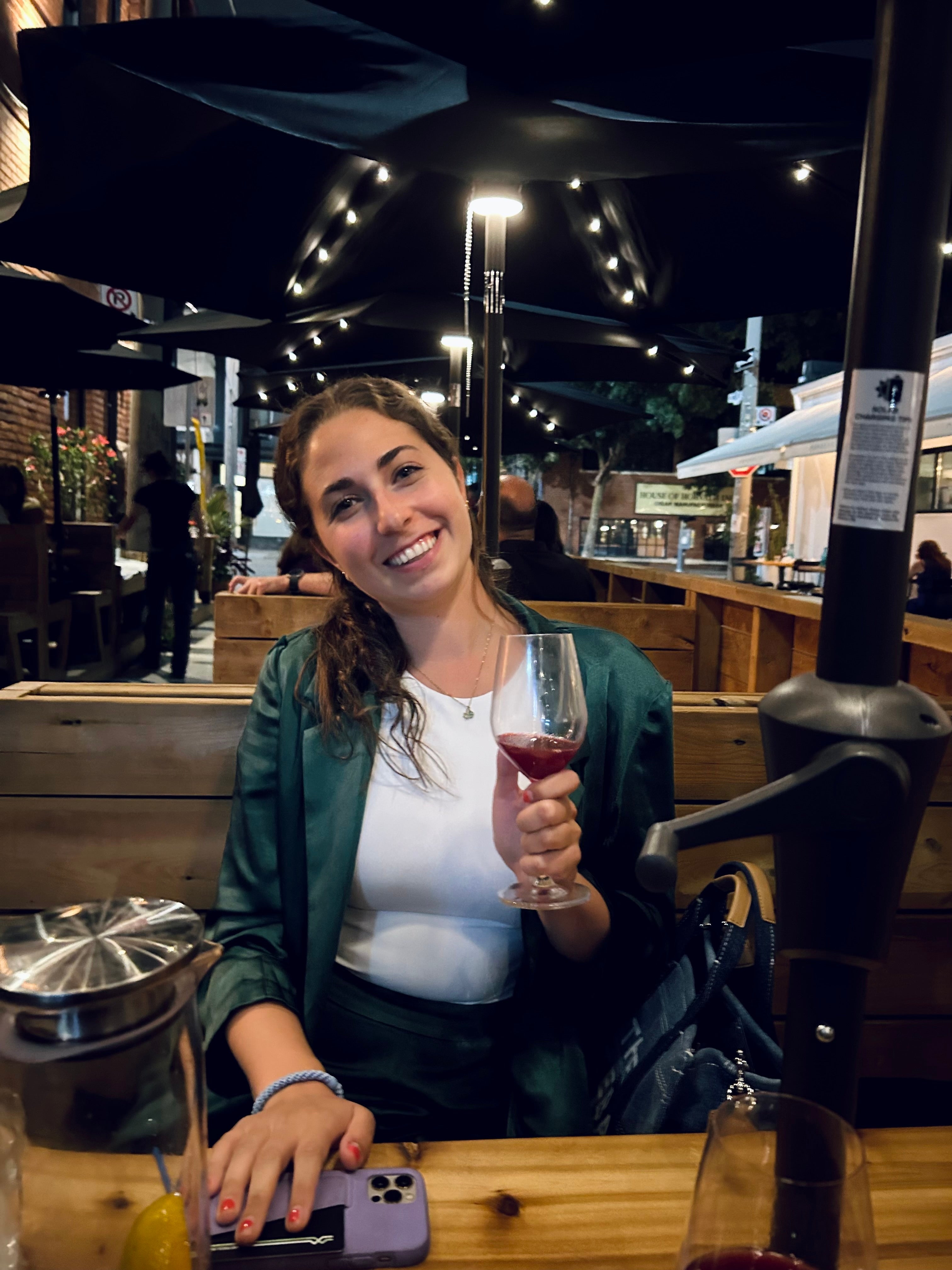Person sits astatine  an outdoor edifice  holding a solid  of wine, smiling, wearing a stylish overgarment   implicit    a casual top