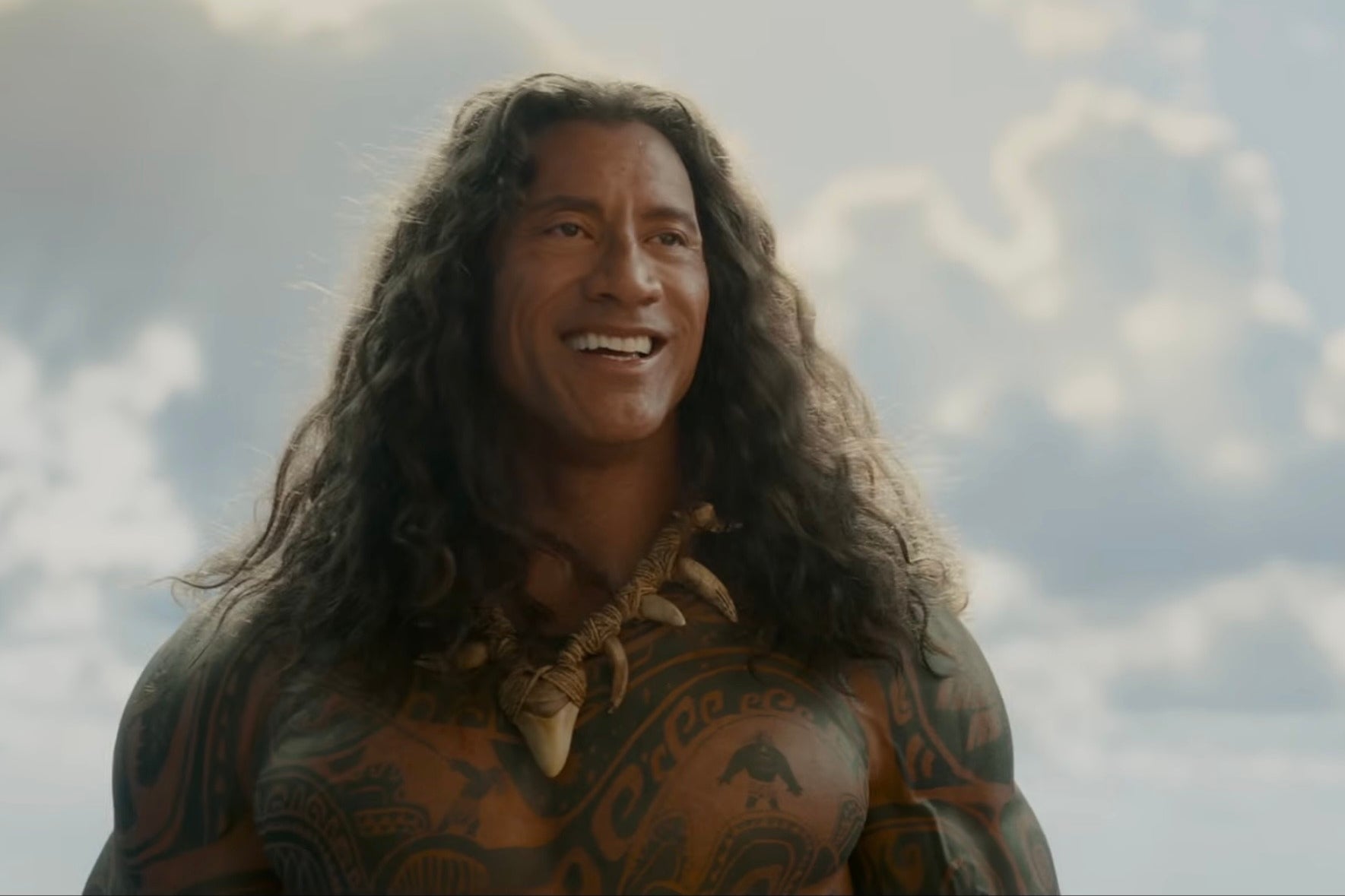Dwayne Johnson with long hair and tribal tattoos stands smiling against a cloudy sky