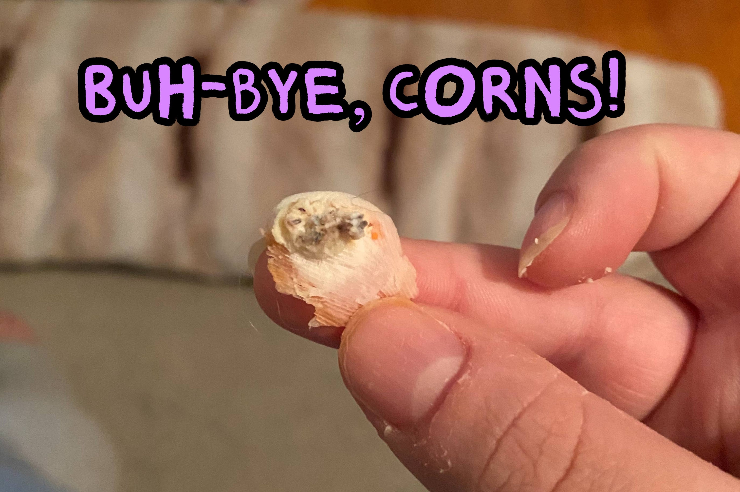 A close-up of a person's hand holding a small removed corn in front of their foot