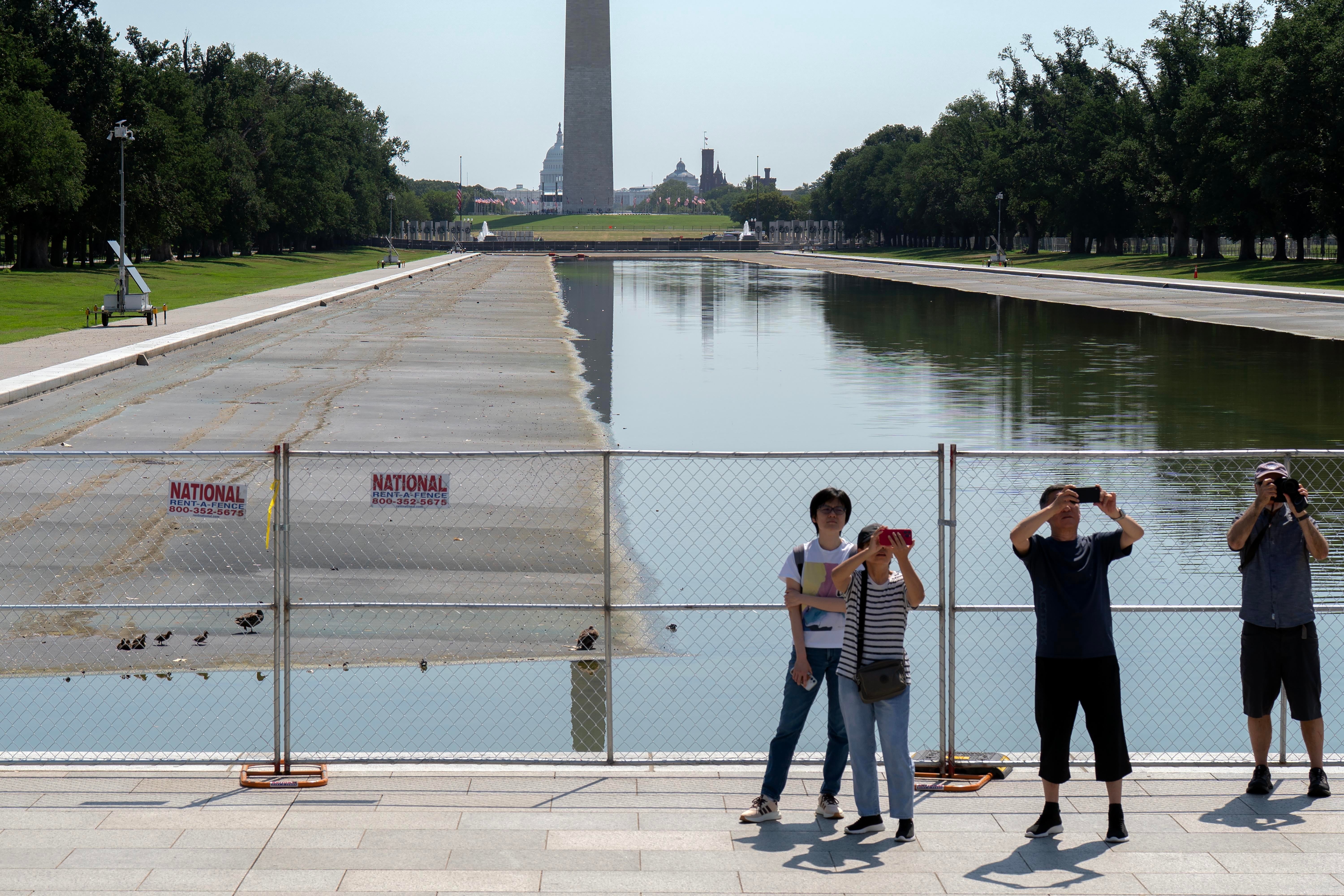 Fire personer står i nærheten av et inngjerdet reflekterende basseng og tar bilder med Washington-monumentet i bakgrunnen