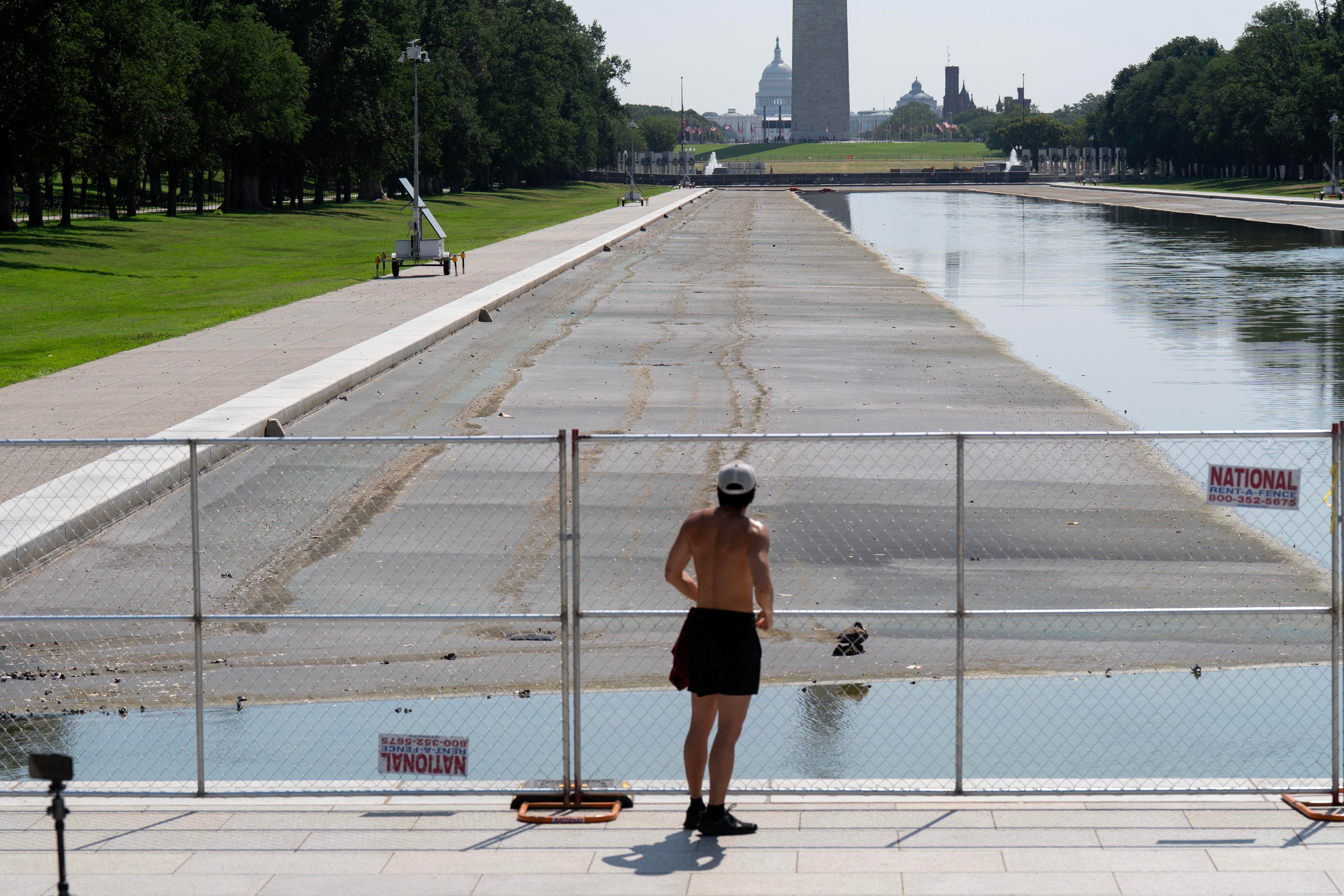 En person i atletisk antrekk står ved et inngjerdet, tomt reflekterende basseng nær et monument i en park