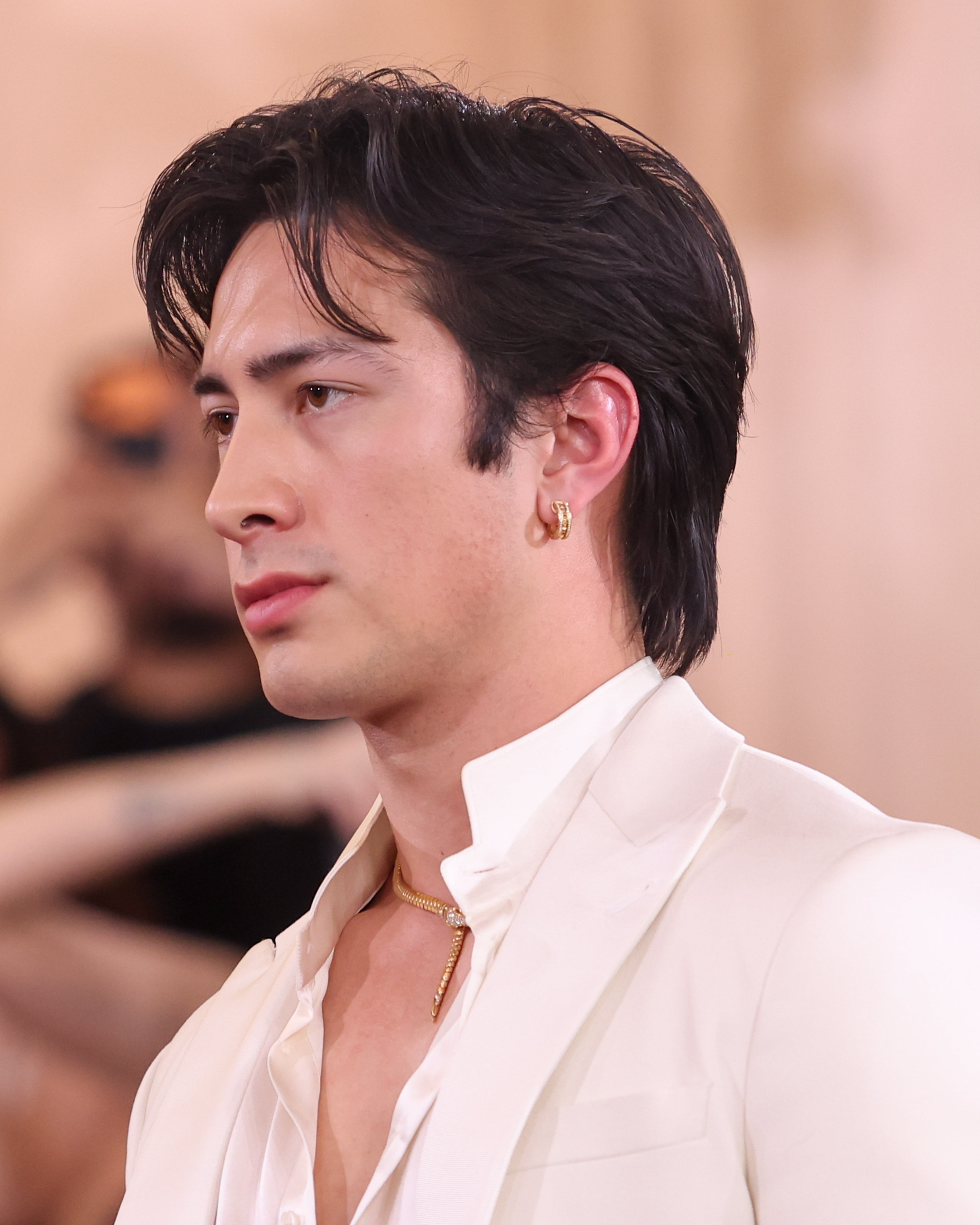 Person in a white suit with gold accessories on a red carpet, looking to the side. Hairstyle is swept back. Audience blurred in the background