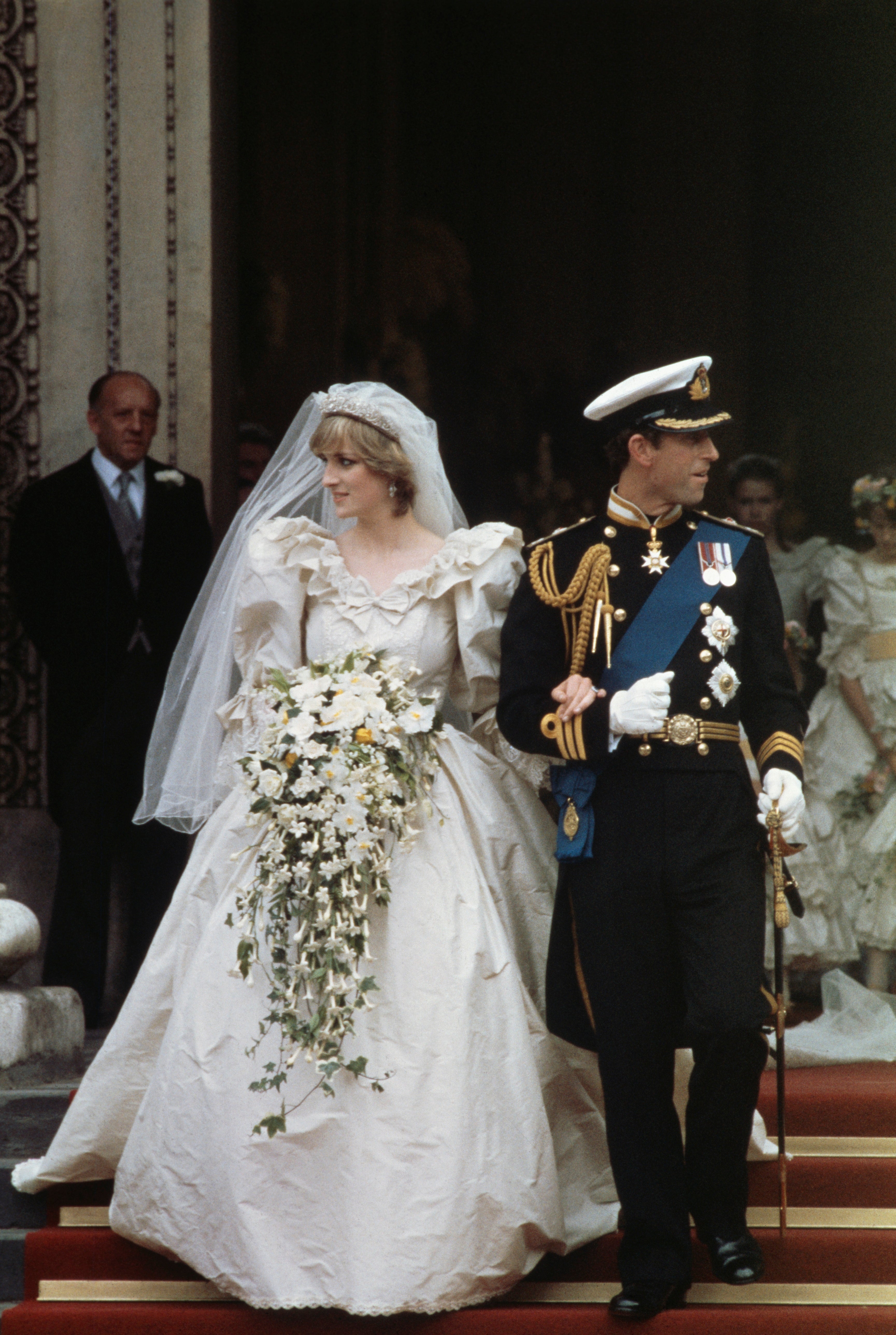A couple in formal attire; the woman wears a ruffled wedding dress with a long veil, the man is in a decorated military uniform