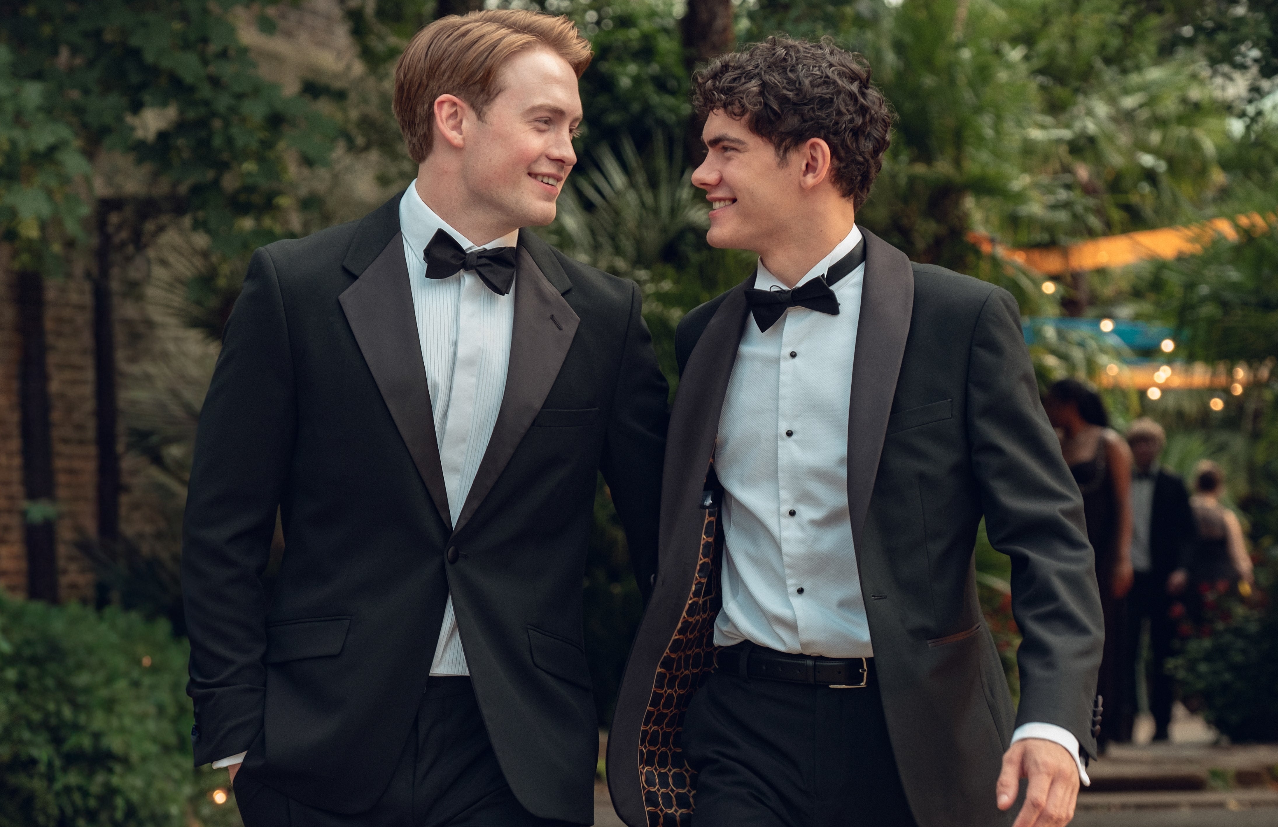 Two people in tuxedos smiling at each other, walking on a garden path