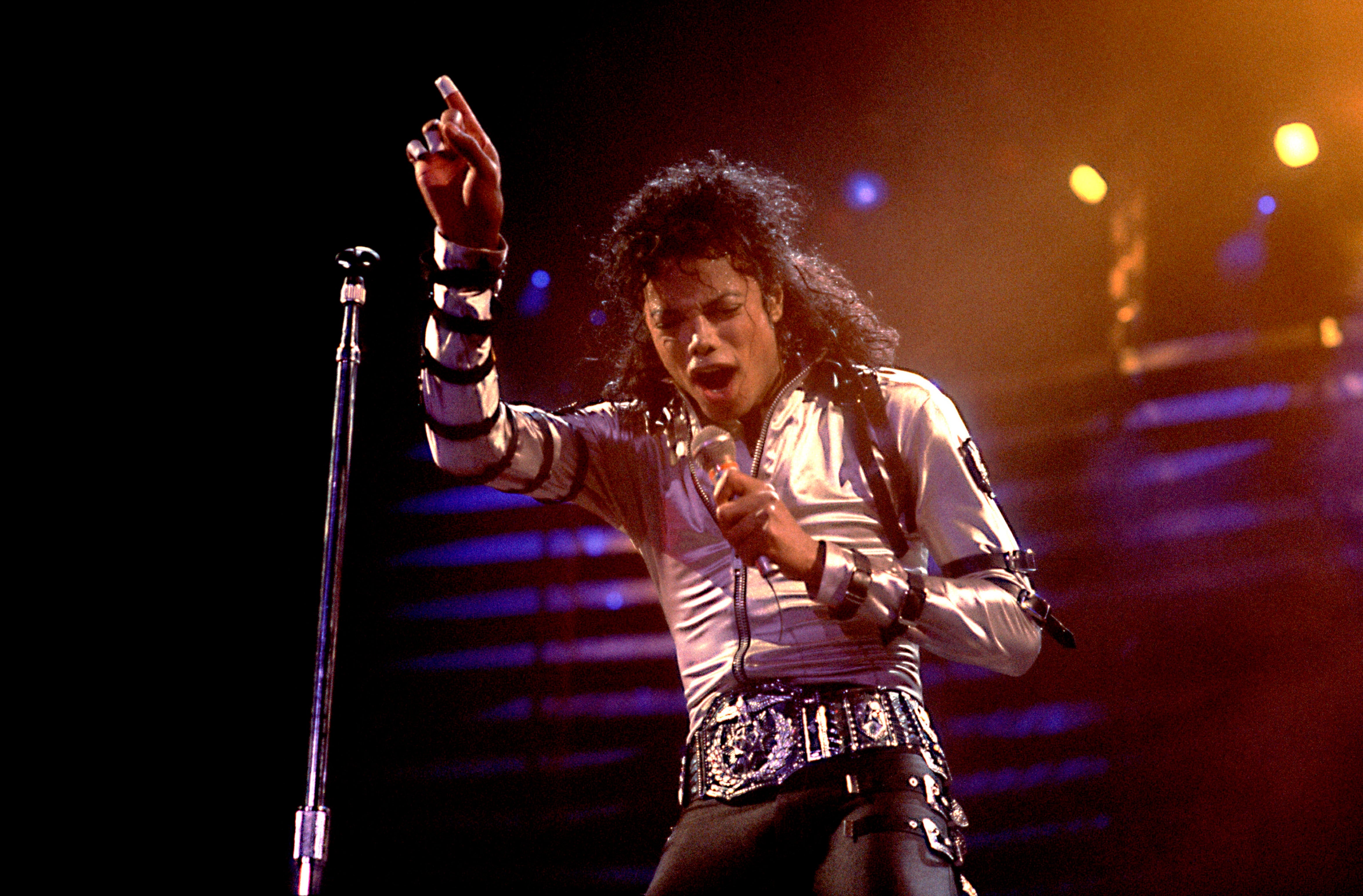 Musician performing energetically on stage, wearing a studded leather jacket and decorative belt, holding a microphone