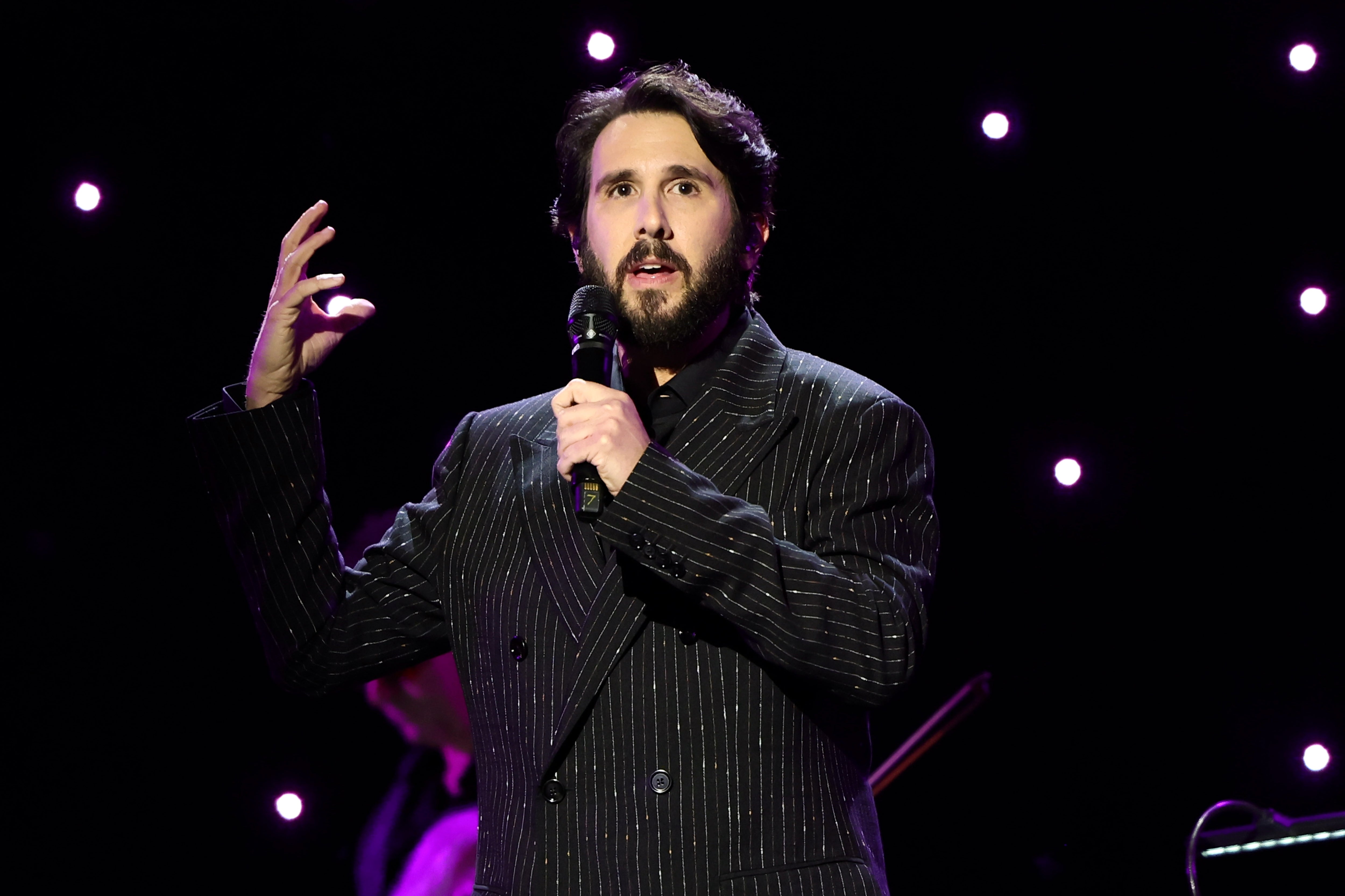A person in a pinstripe suit is performing on stage, holding a microphone, with a starry backdrop