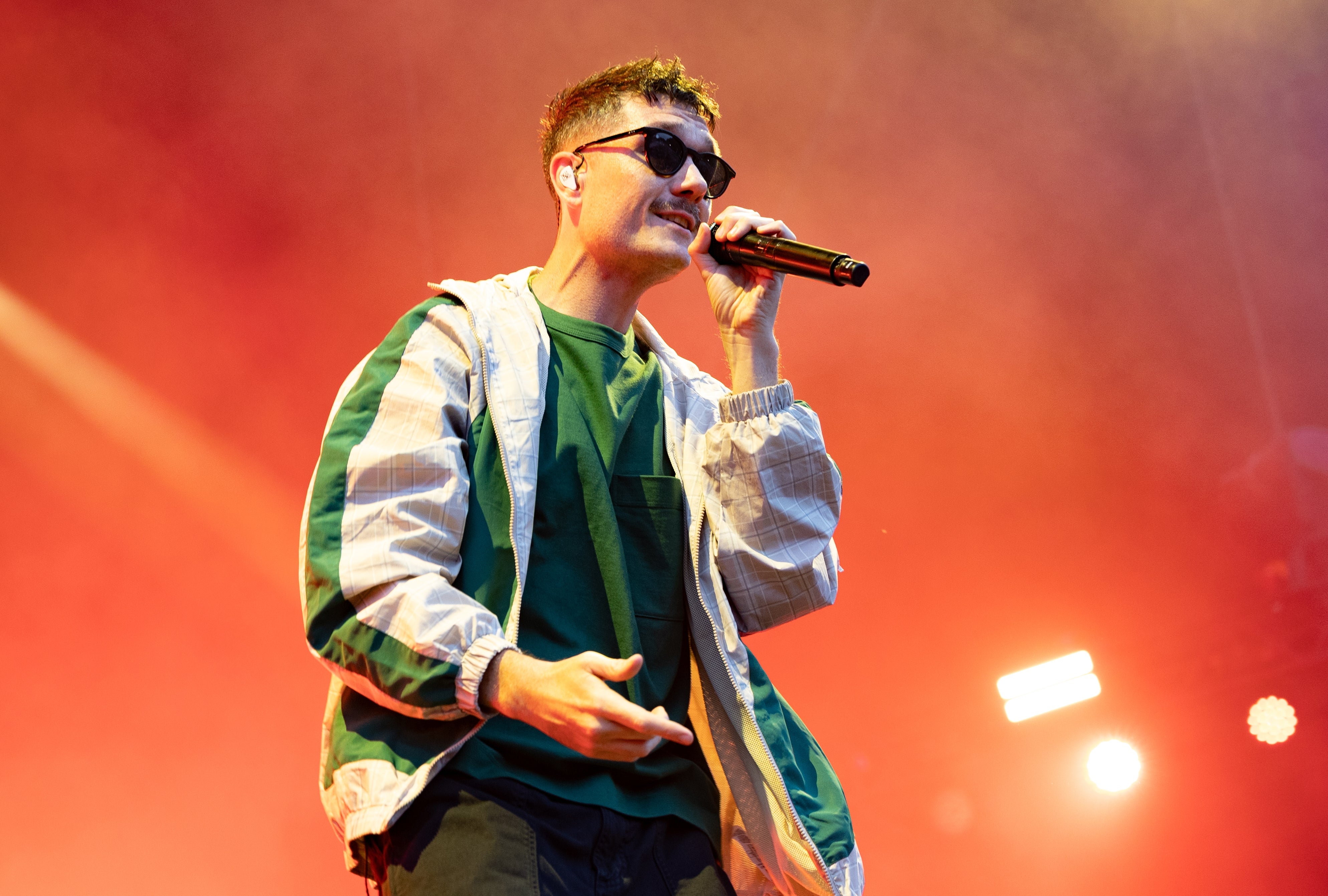 Performer on stage in casual outfit, wearing sunglasses, holding microphone mid-performance against a vibrant background