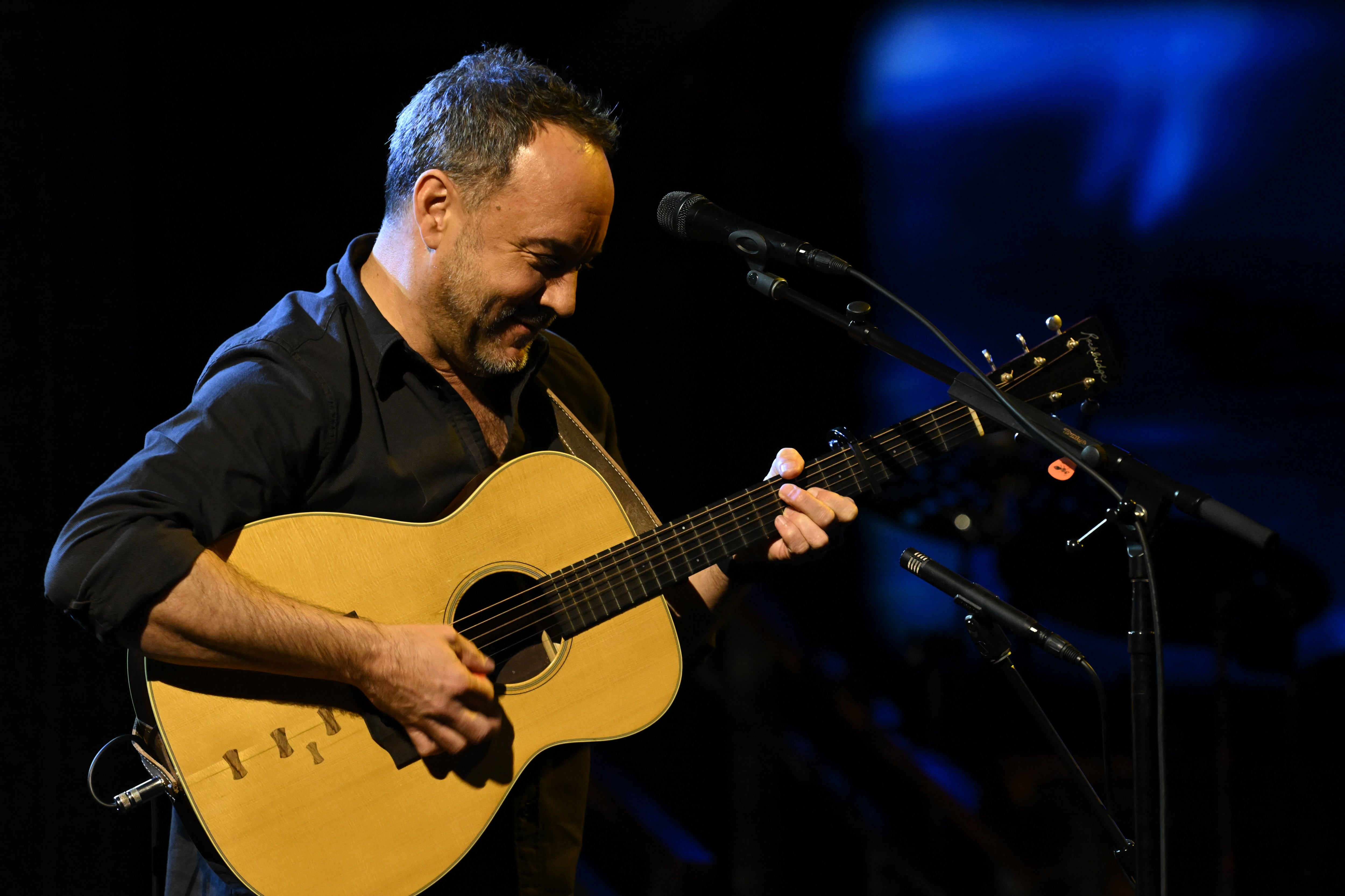 Musician playing an acoustic guitar on stage. Wearing a black shirt, he is focused on his performance, surrounded by microphones