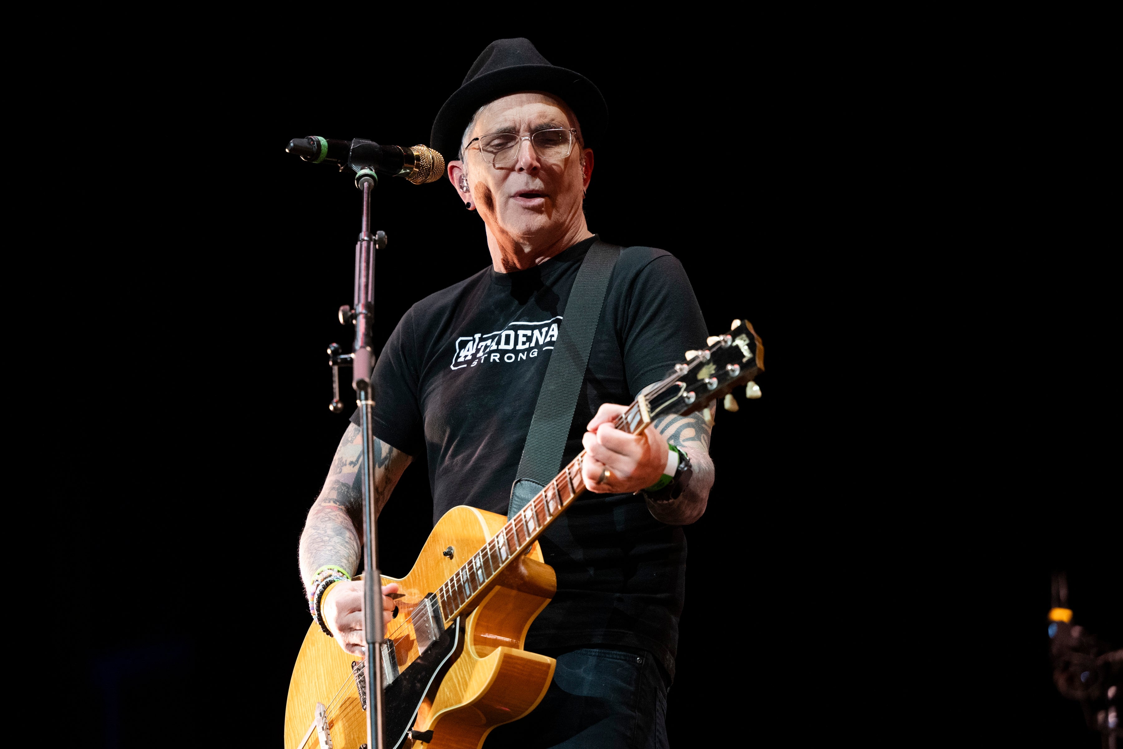 Musician playing guitar on stage, wearing a fedora and t-shirt, performing energetically while singing into a microphone