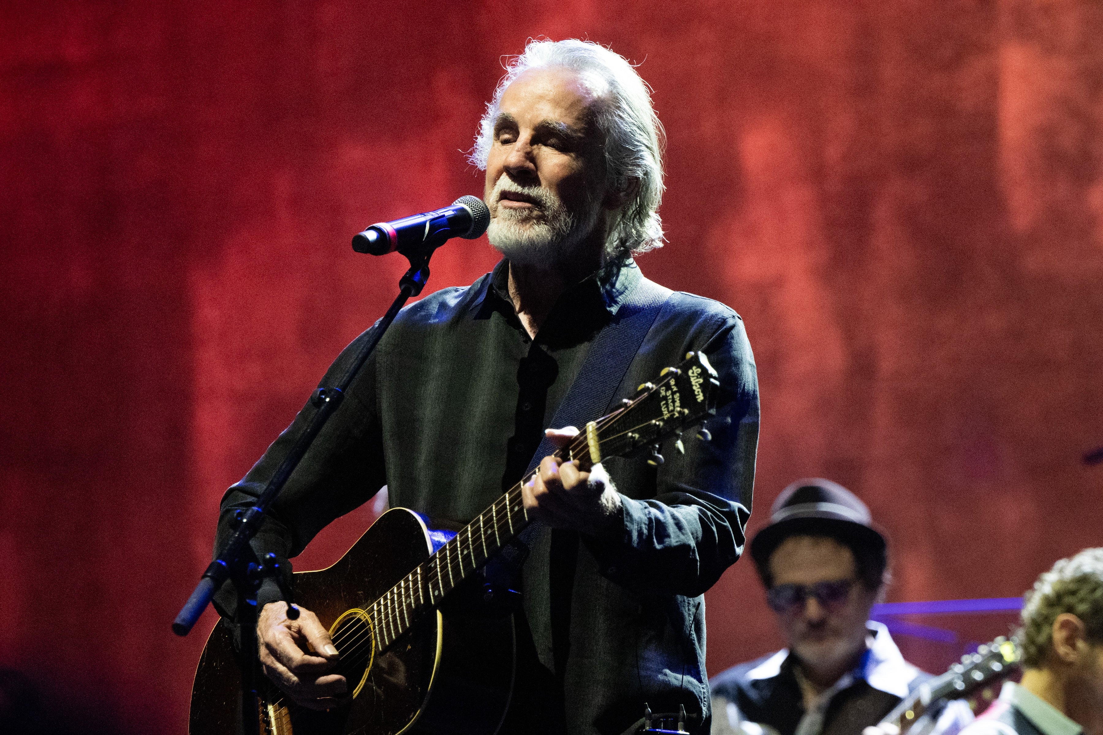 A musician performs on stage, playing an acoustic guitar and singing into a microphone. His style is classic and relaxed