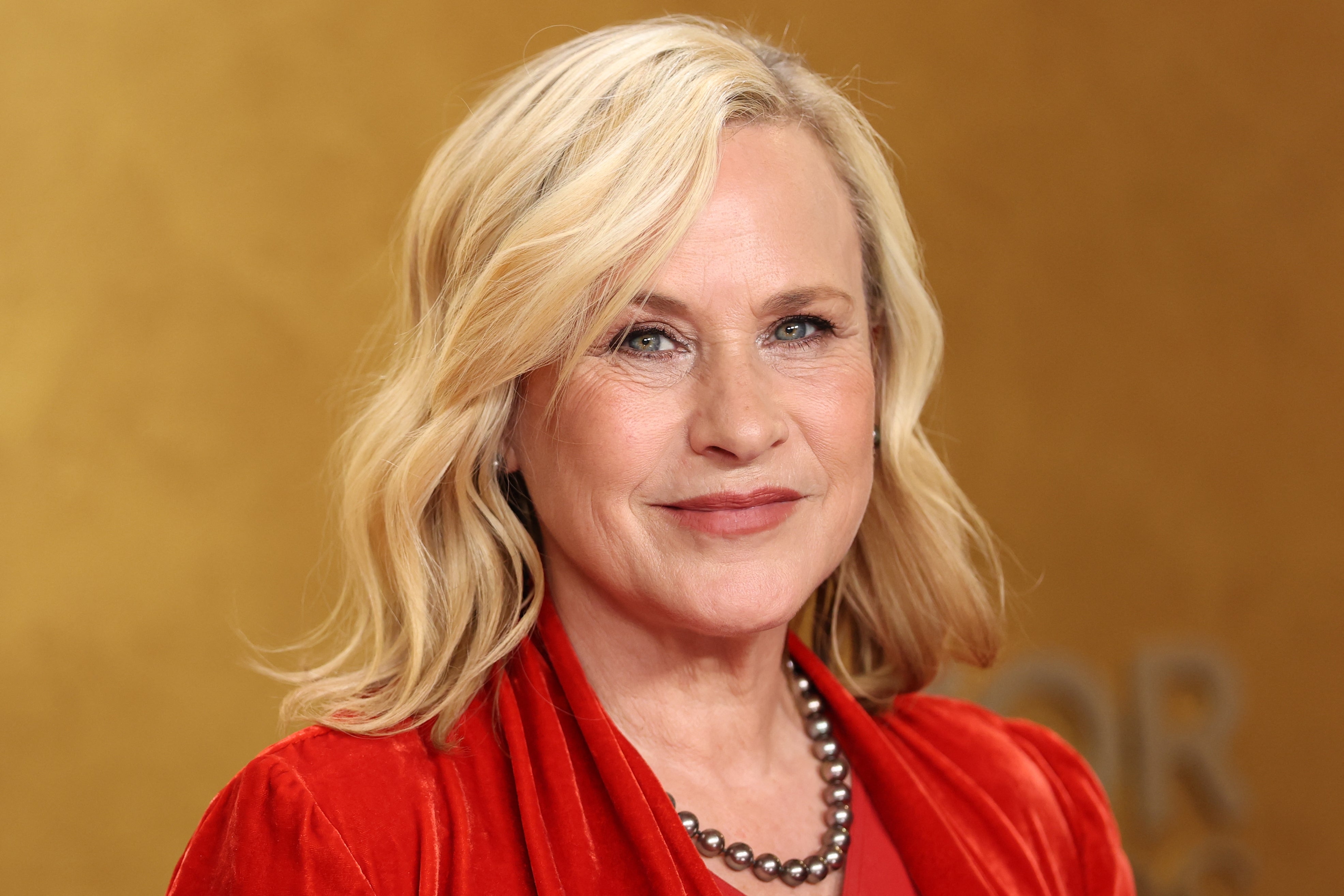 A person with shoulder-length hair, wearing a red velvet dress and a pearl necklace, smiles at a celebrity event