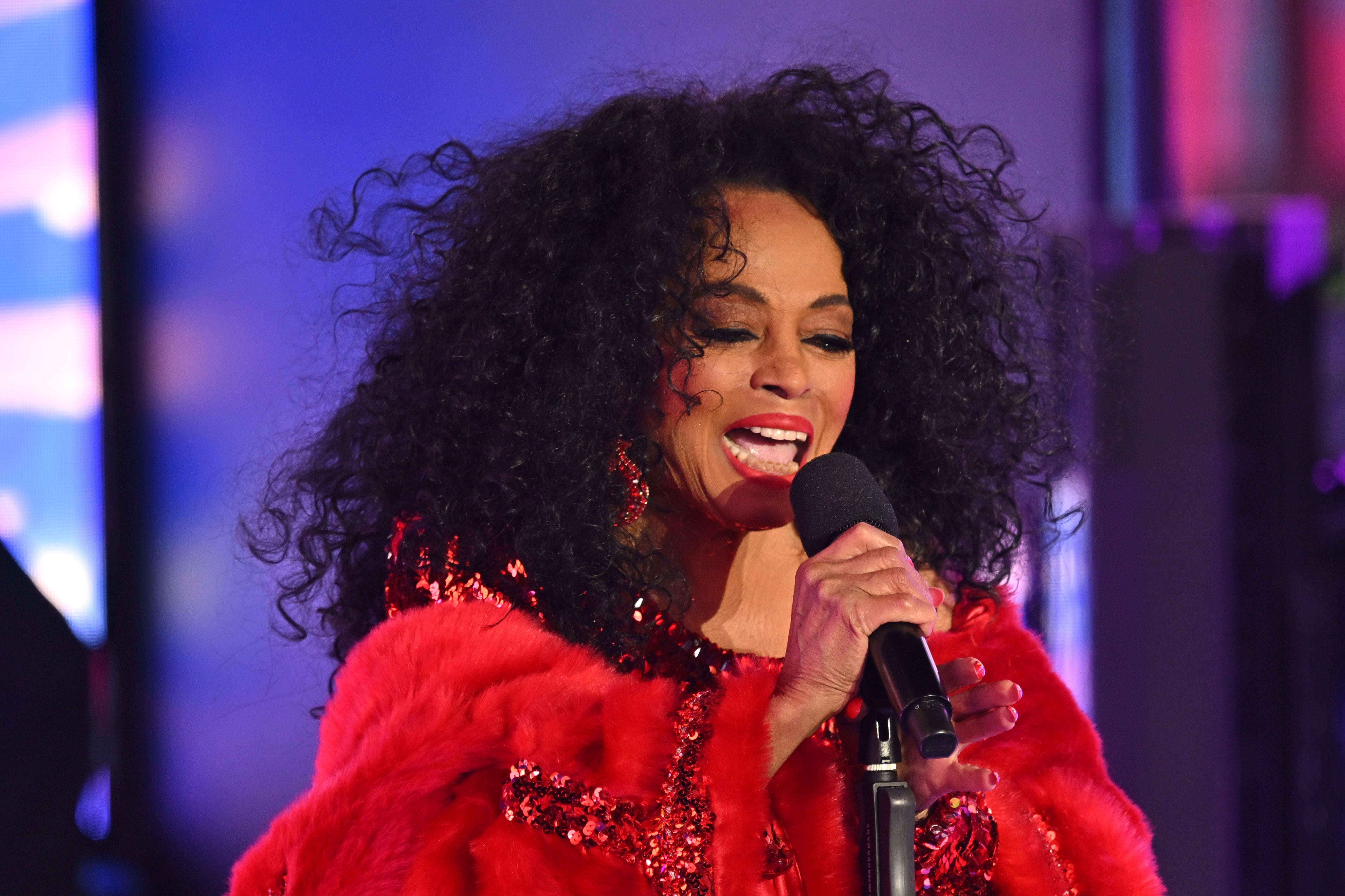 Singer performing on stage, wearing a sparkling, patterned outfit with a fluffy wrap, holding a microphone
