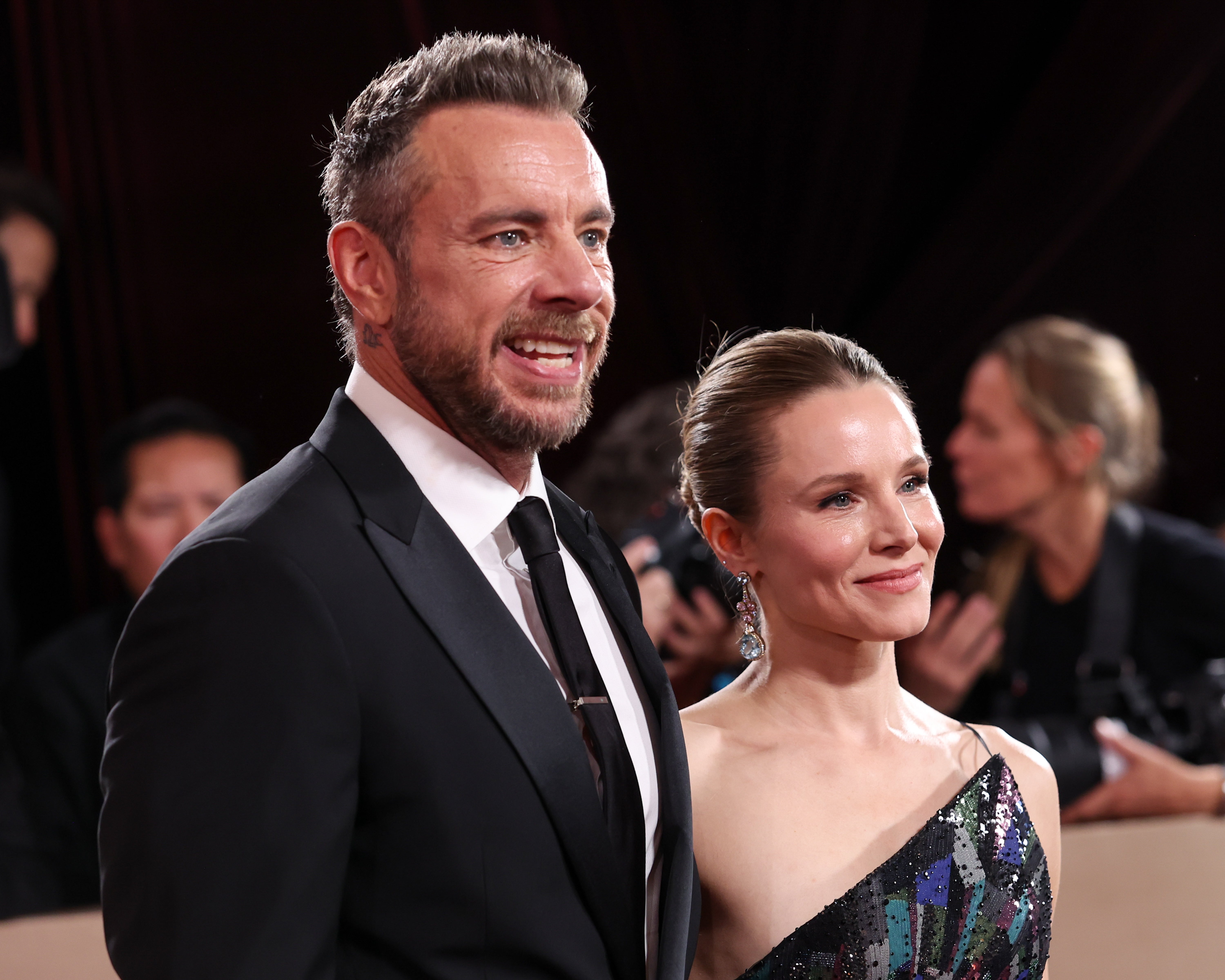 A man in a classic tuxedo and a woman in an asymmetrical sequined gown smile on the red carpet. Photographers are visible in the background