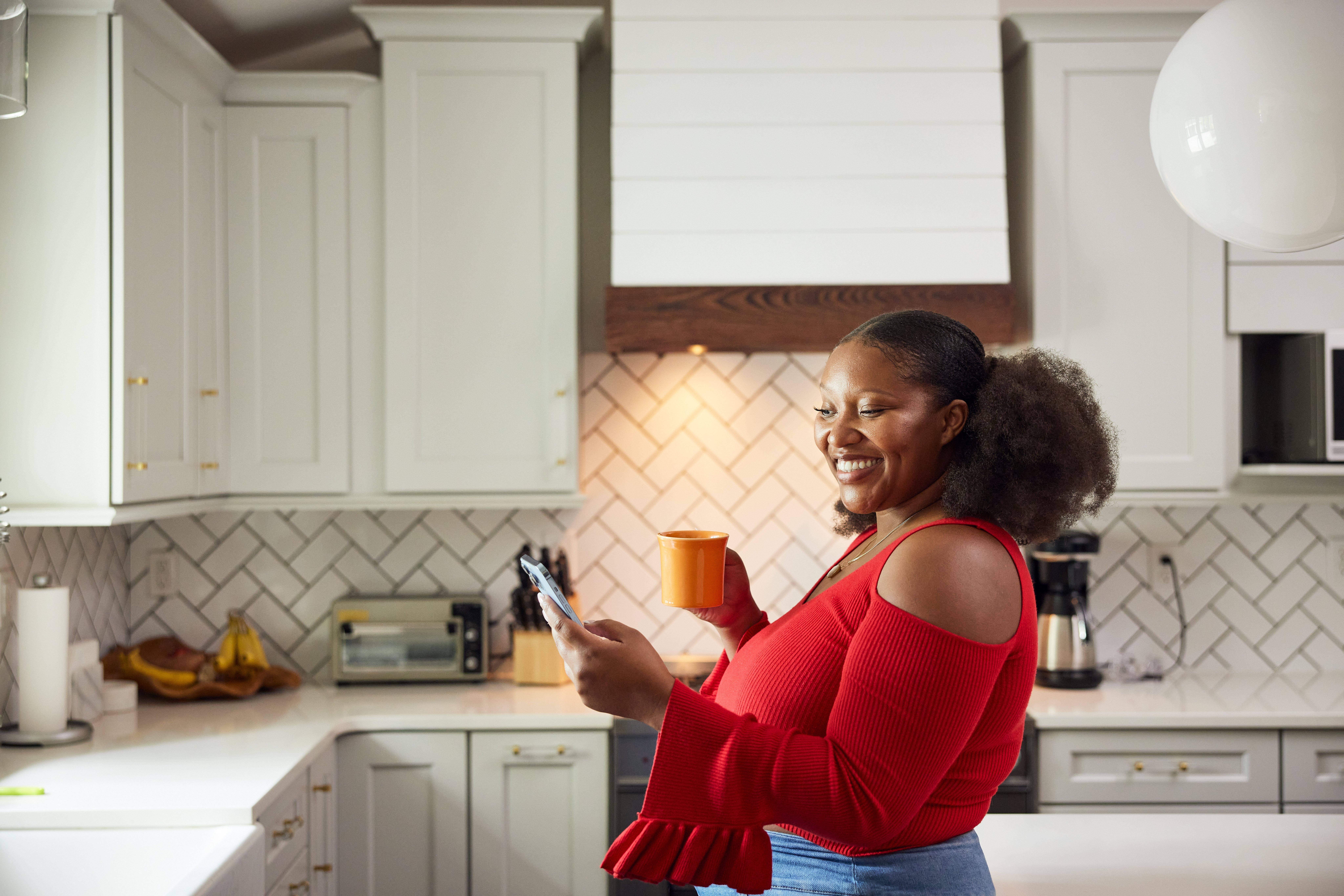 Person successful  stylish reddish  top, smiling portion    holding a telephone  and a mug successful  a modern   kitchen