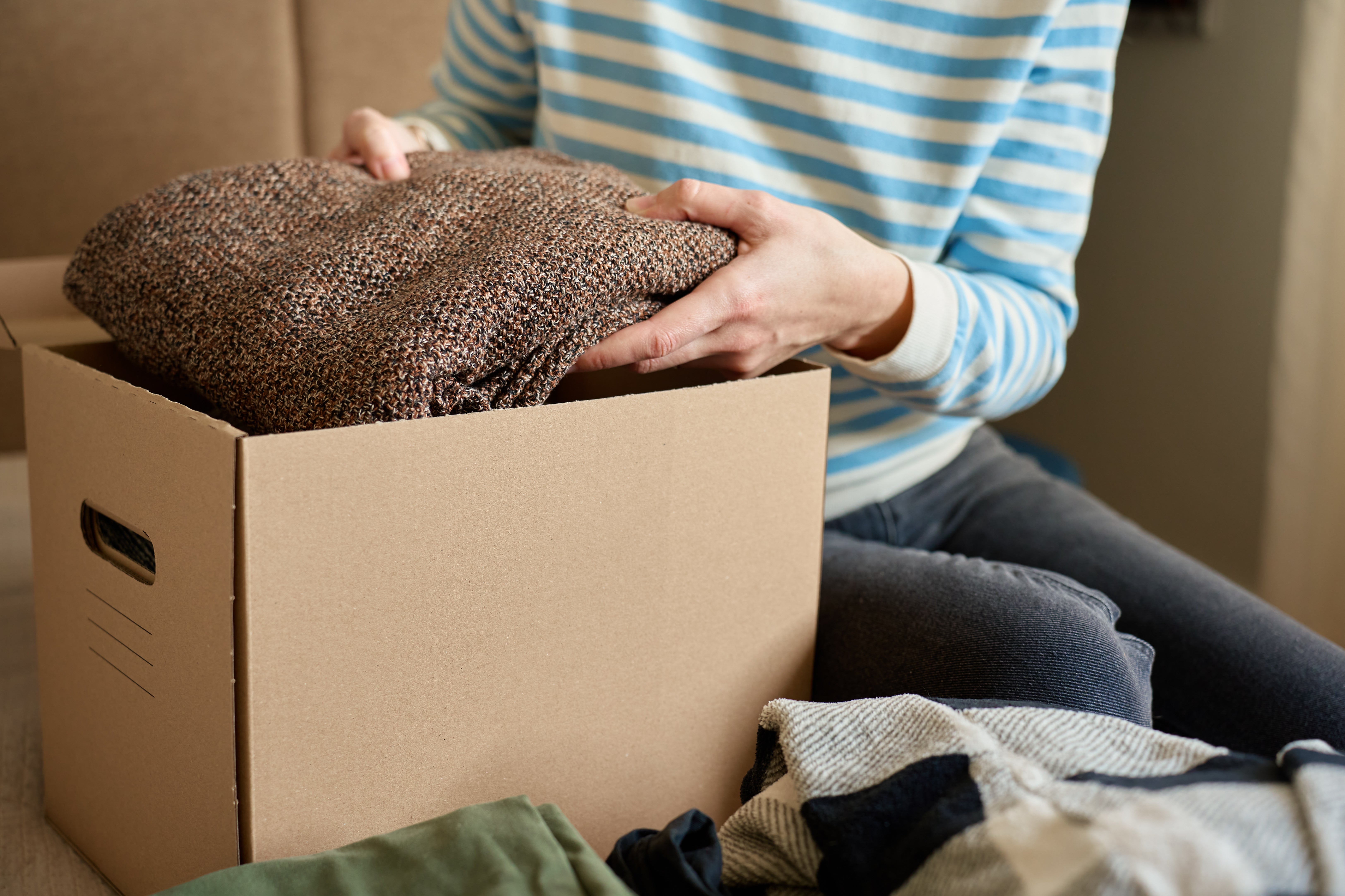 Person successful striped garment packing a cardboard container with clothes; nearby, folded garments are visible